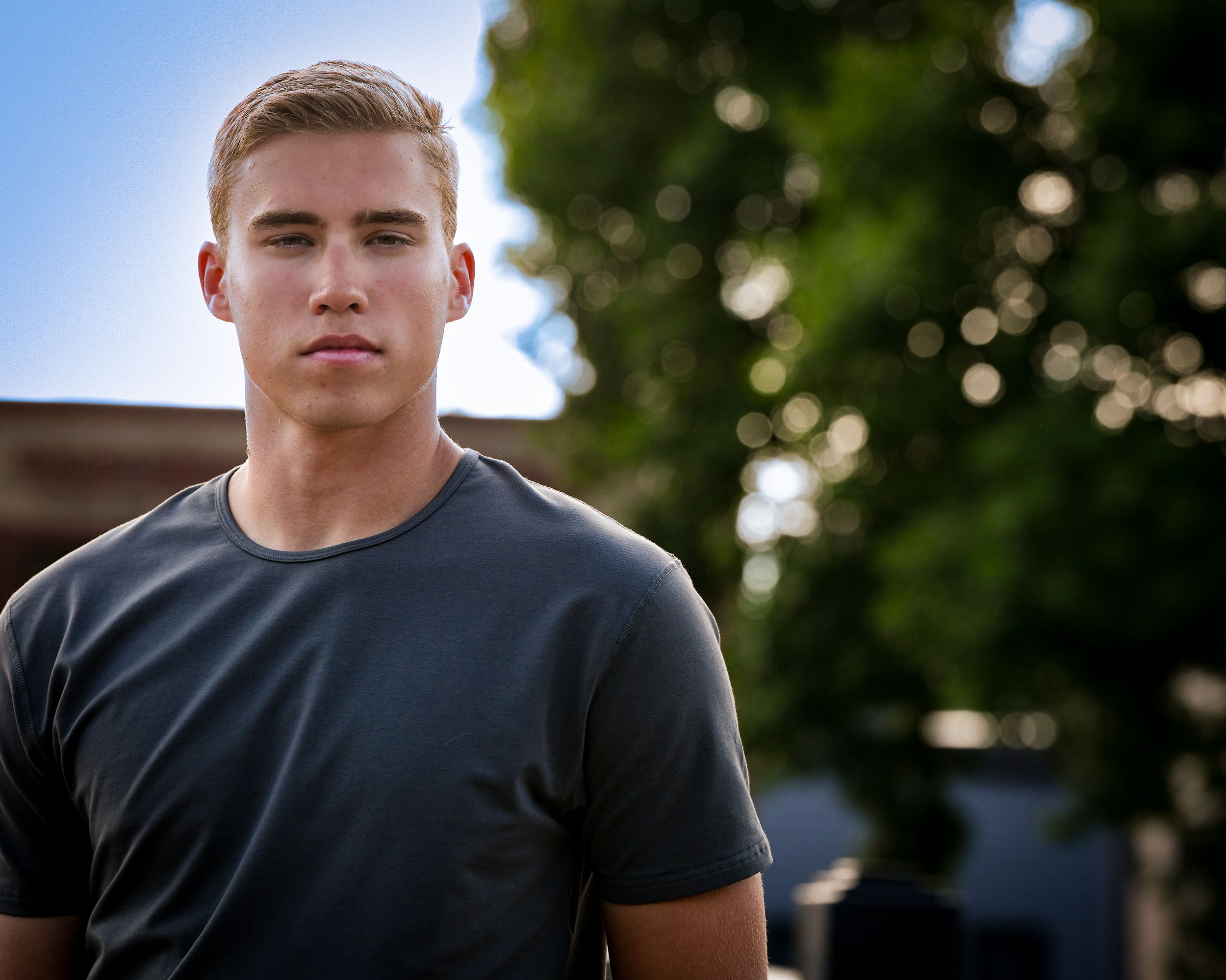 Young blond man in dark gray t-shirt posing outdoors with green trees in blurred background.