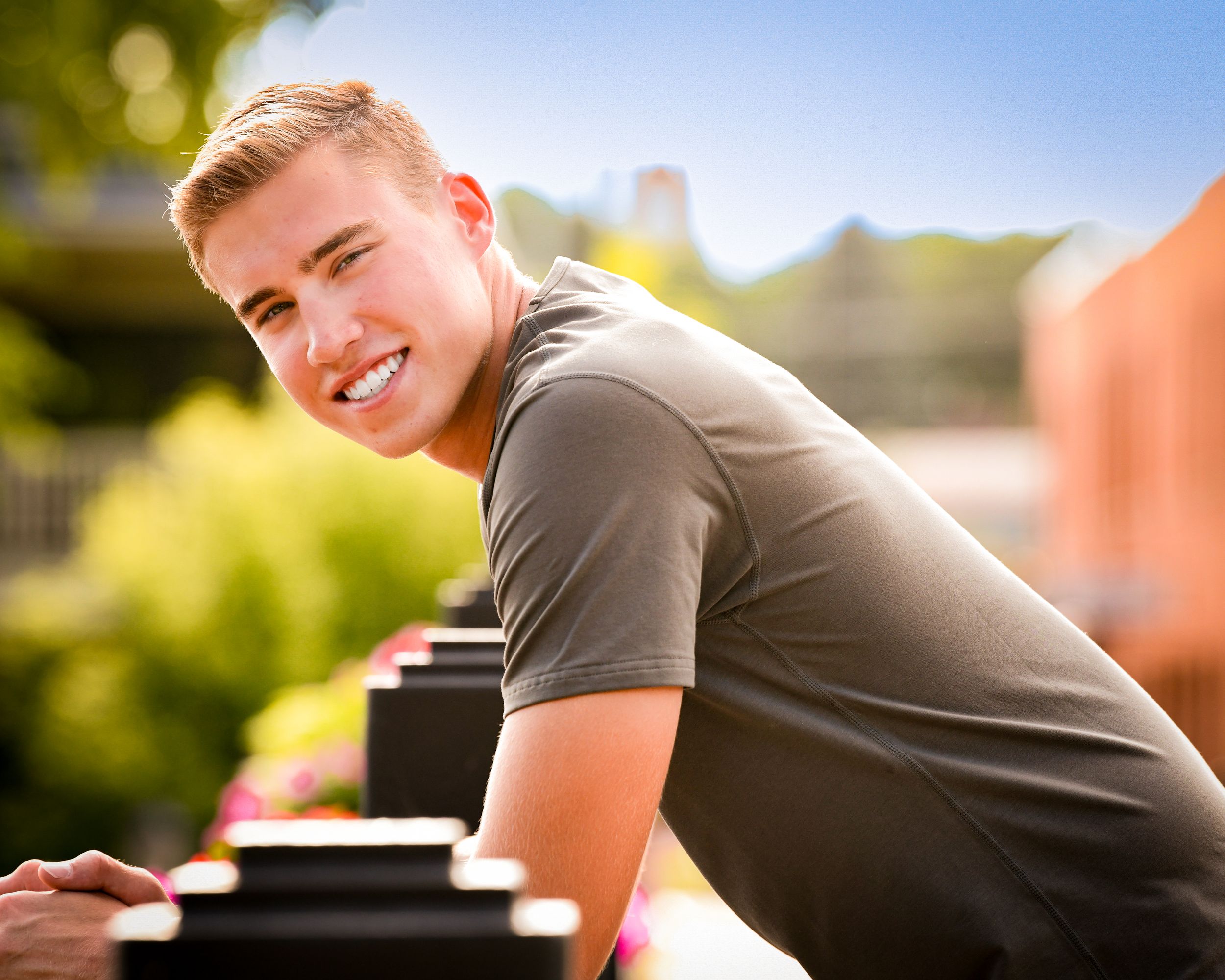 Smiling young man in gray t-shirt leaning on railing outdoors on a sunny day.