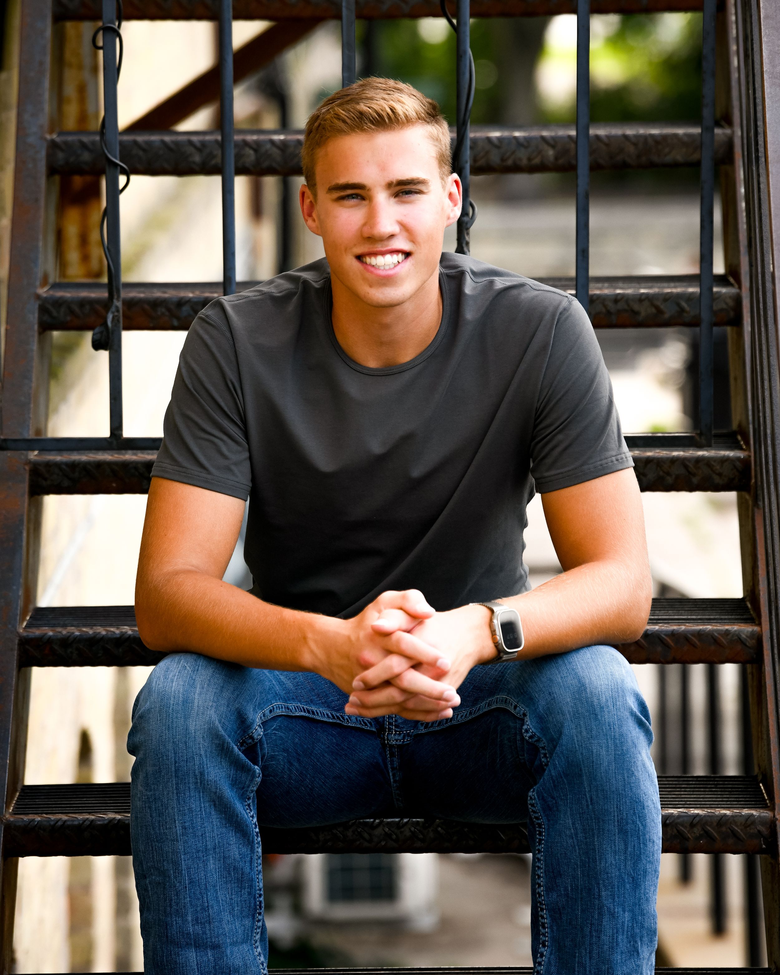 Smiling young man in gray t-shirt and jeans sitting on metal stairs outdoors.