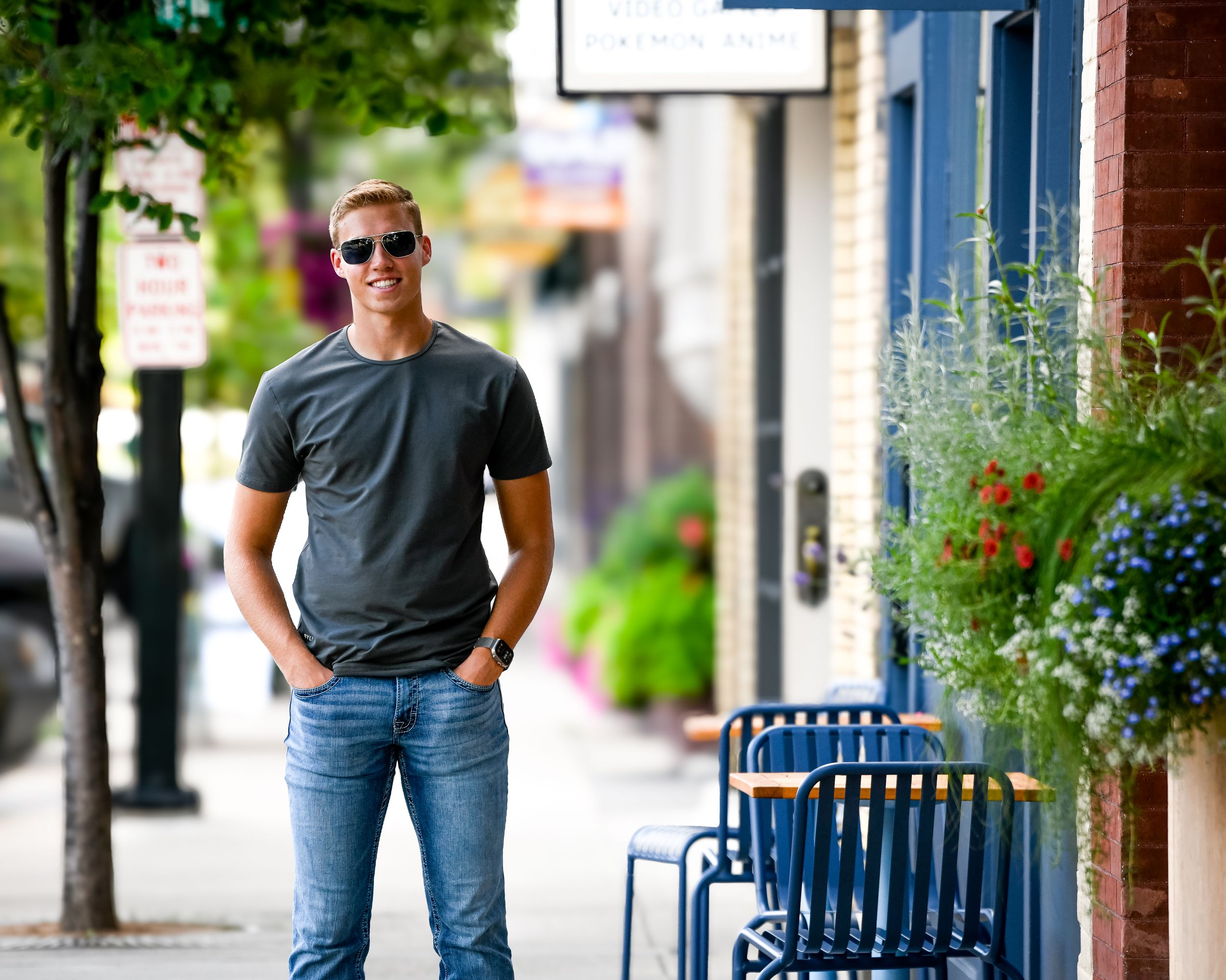 Young man in gray t-shirt and jeans with sunglasses standing on a sunny urban sidewalk near shops.