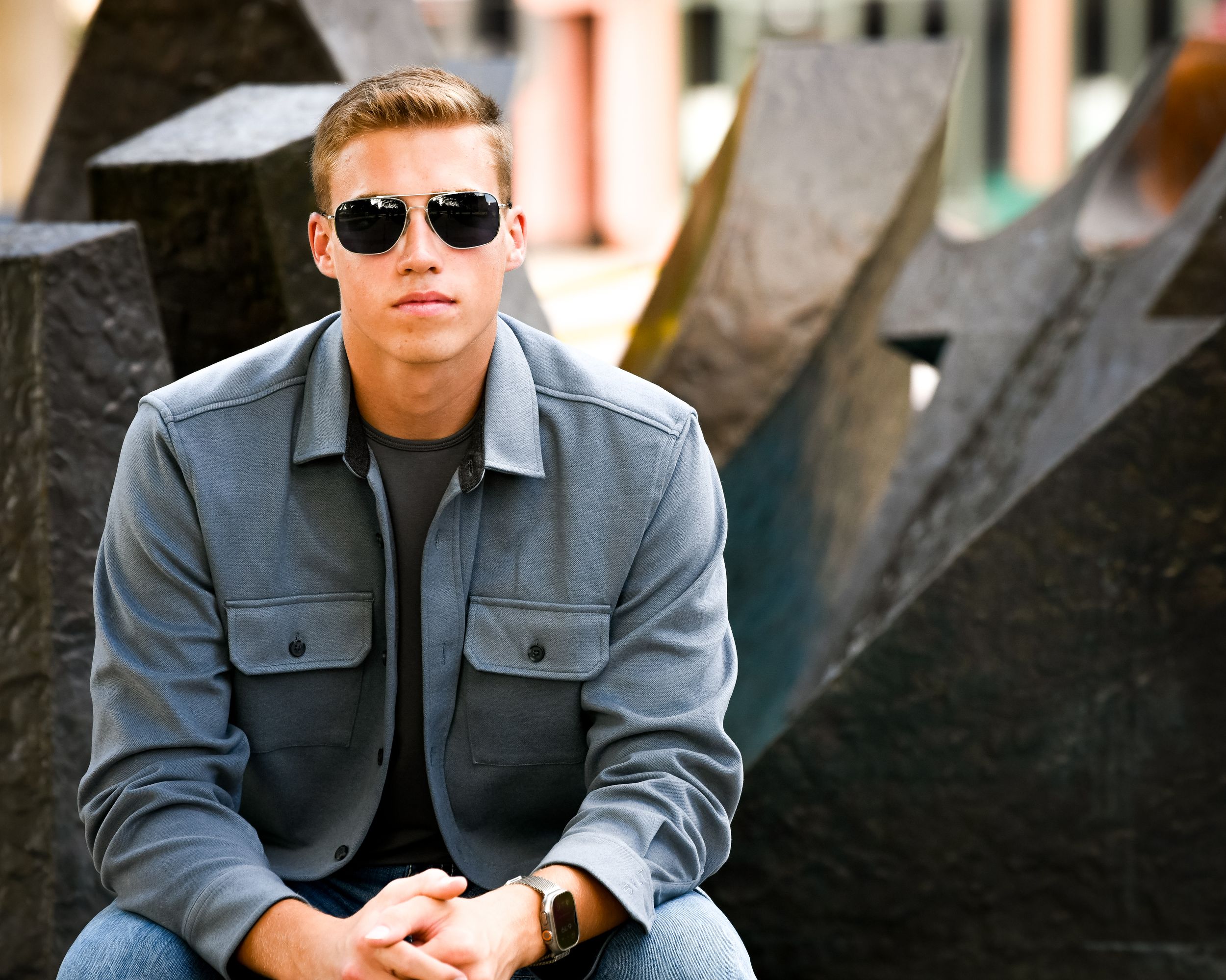 Young man in gray jacket and sunglasses sitting outdoors near abstract metal sculpture.