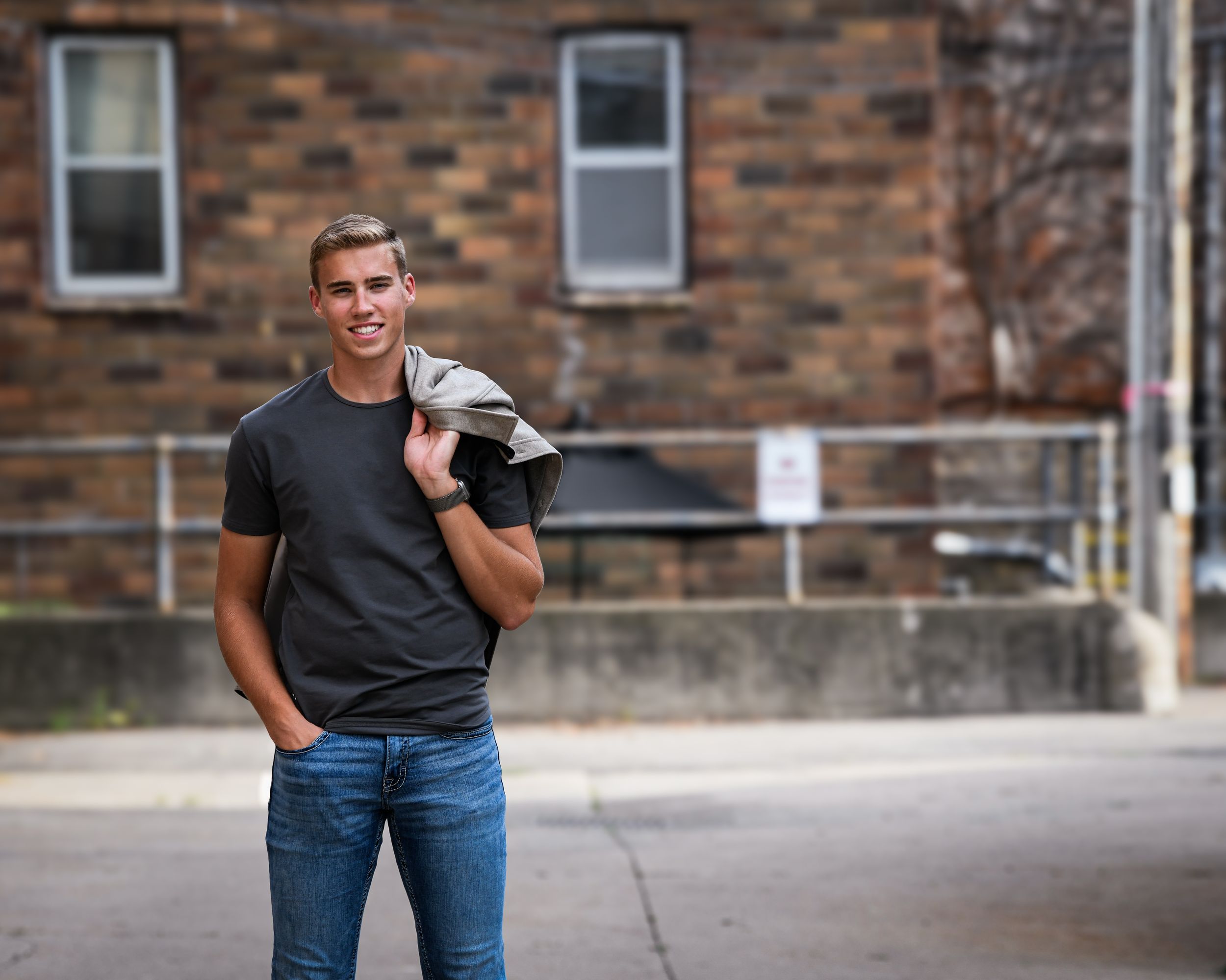 Young man in black t-shirt and jeans holding jacket over shoulder, smiling outdoors near brick building.