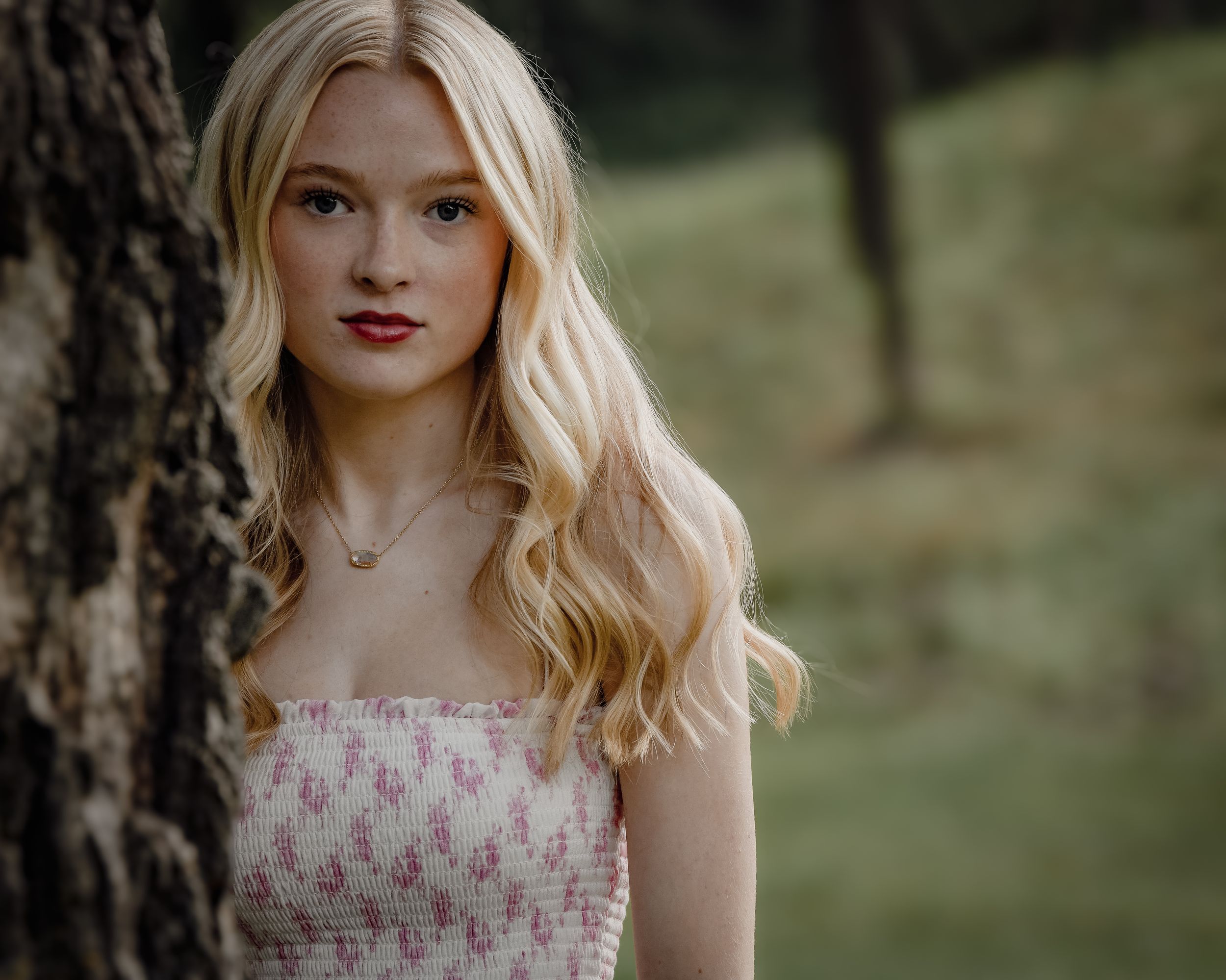 Young blonde woman in pink floral dress posing near tree trunk in outdoor portrait photography.