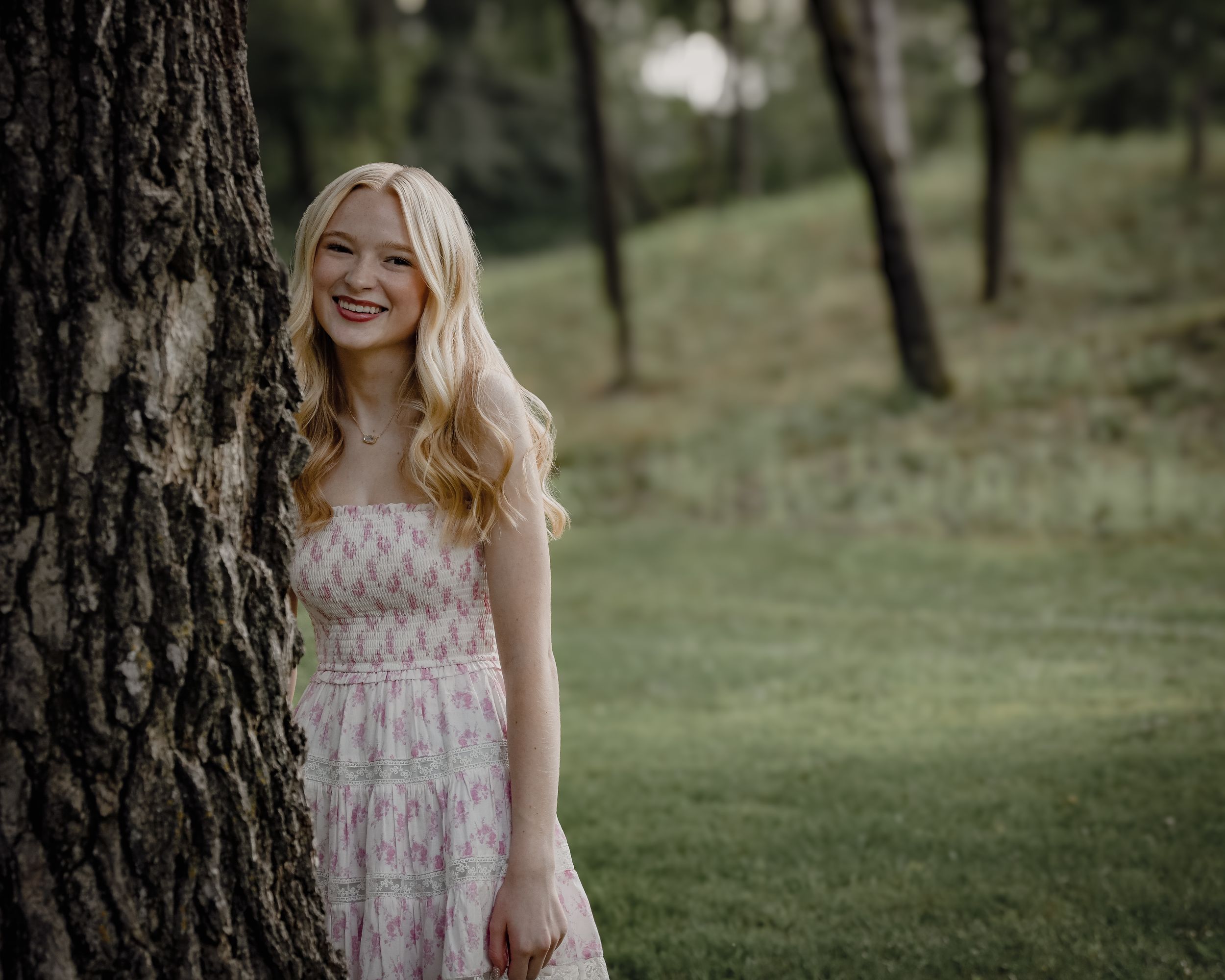 Young blonde woman in floral dress smiling beside a tree in a lush green park.
