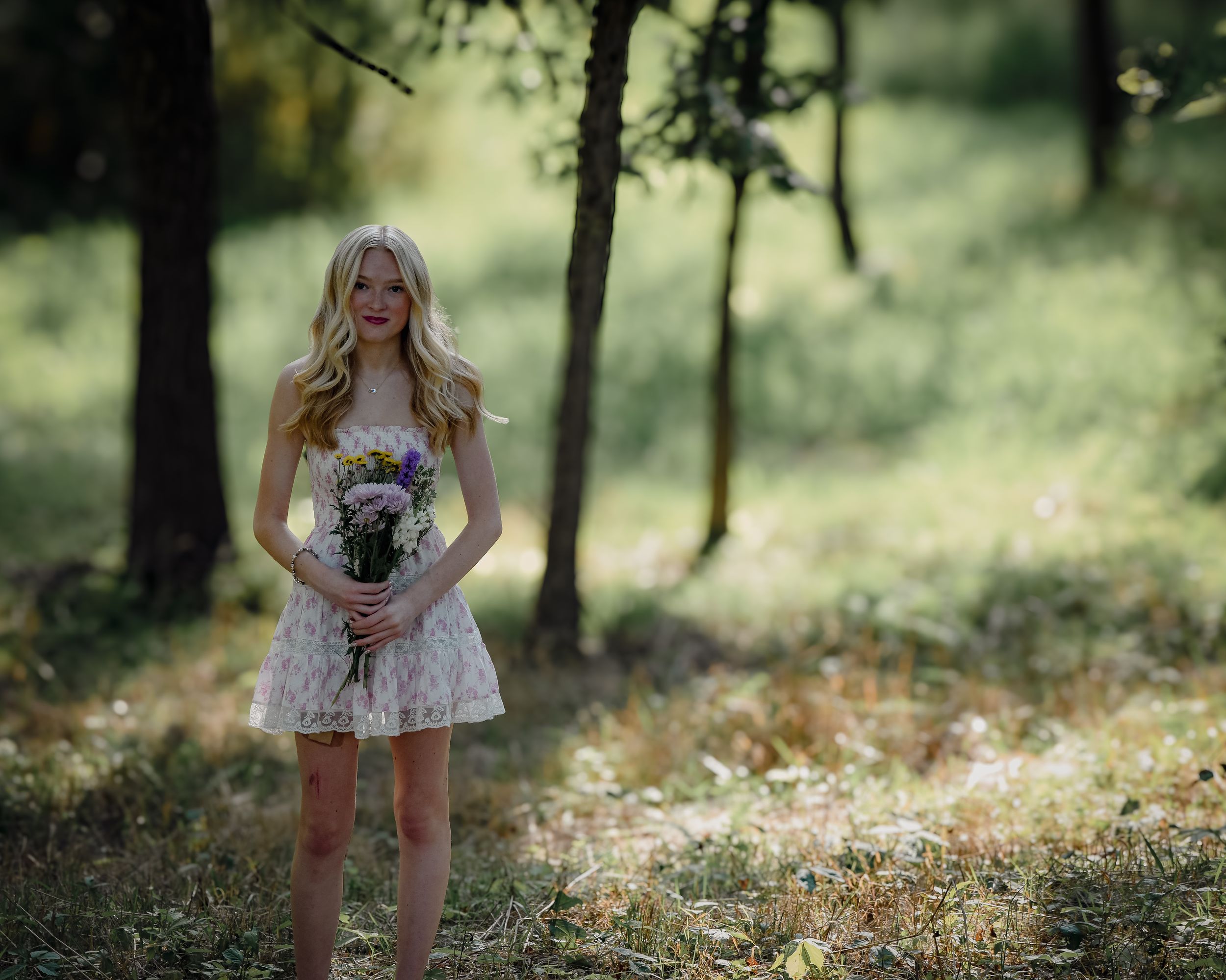 Blonde woman in white dress holding wildflower bouquet smiling in sunlit forest clearing.