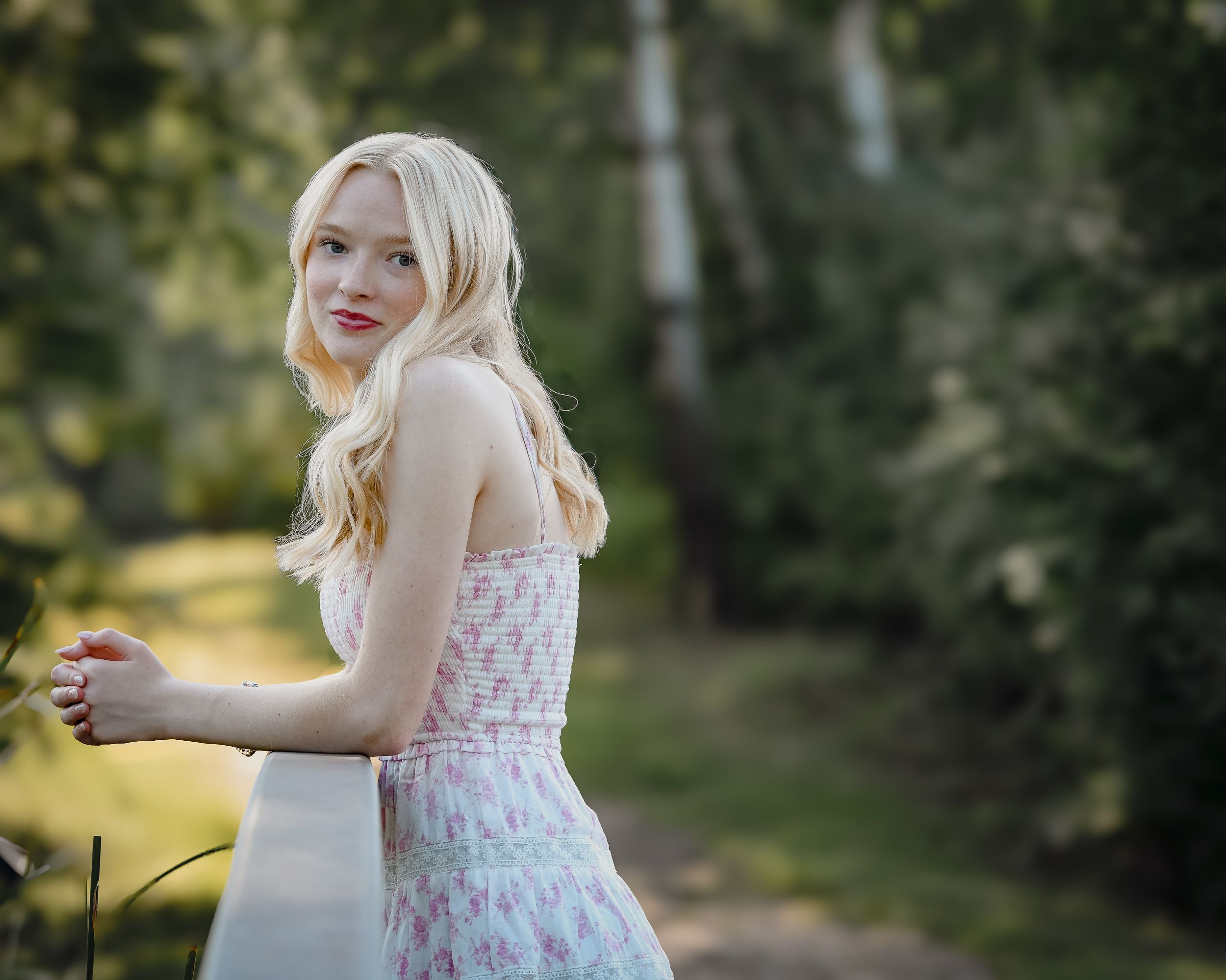 Young blonde woman in floral sundress leaning on white fence in green garden setting.