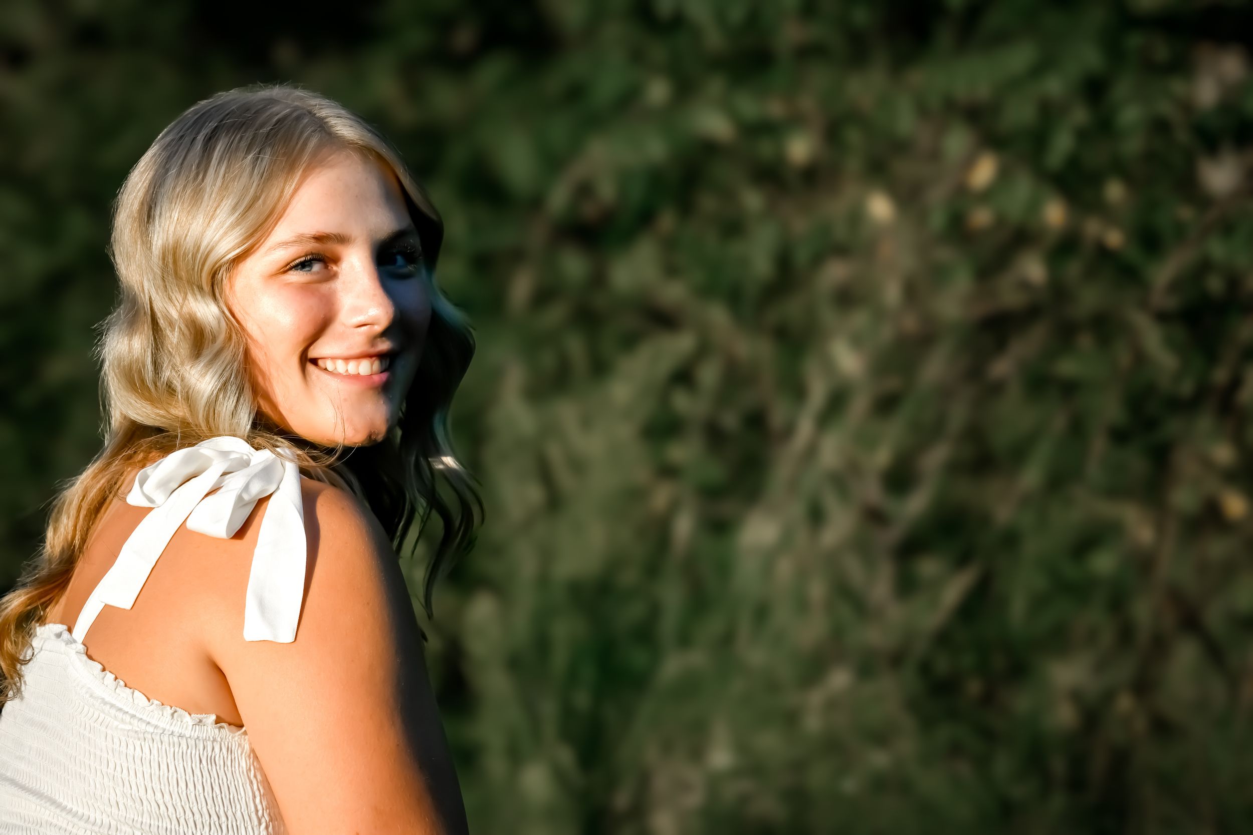 Smiling blonde woman in white dress with tie shoulders, glancing back over her shoulder outdoors.