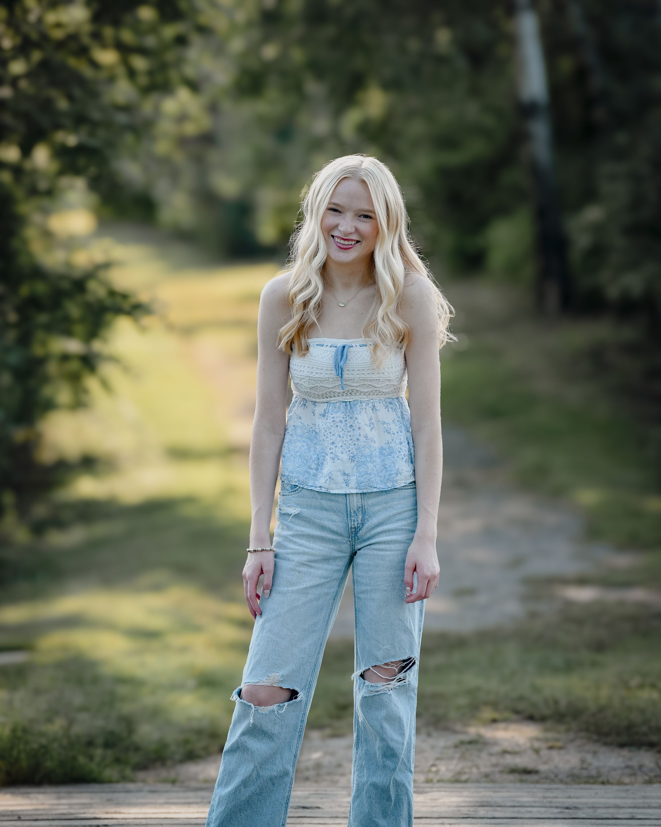 Smiling young woman with blonde hair wearing denim overalls and ripped jeans standing on a path outdoors.