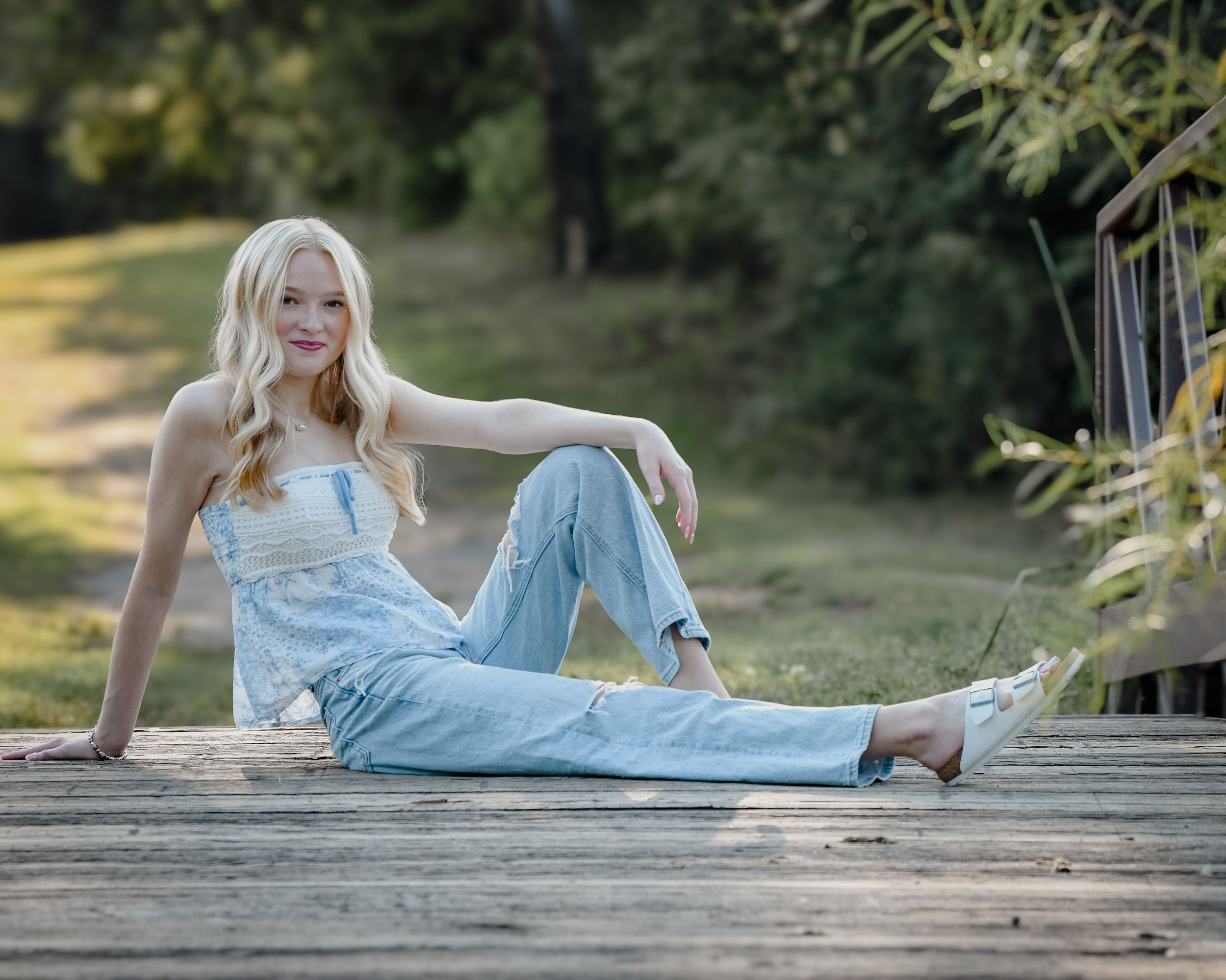 Young blonde woman in white overalls sitting relaxed on wooden deck in sunny park.