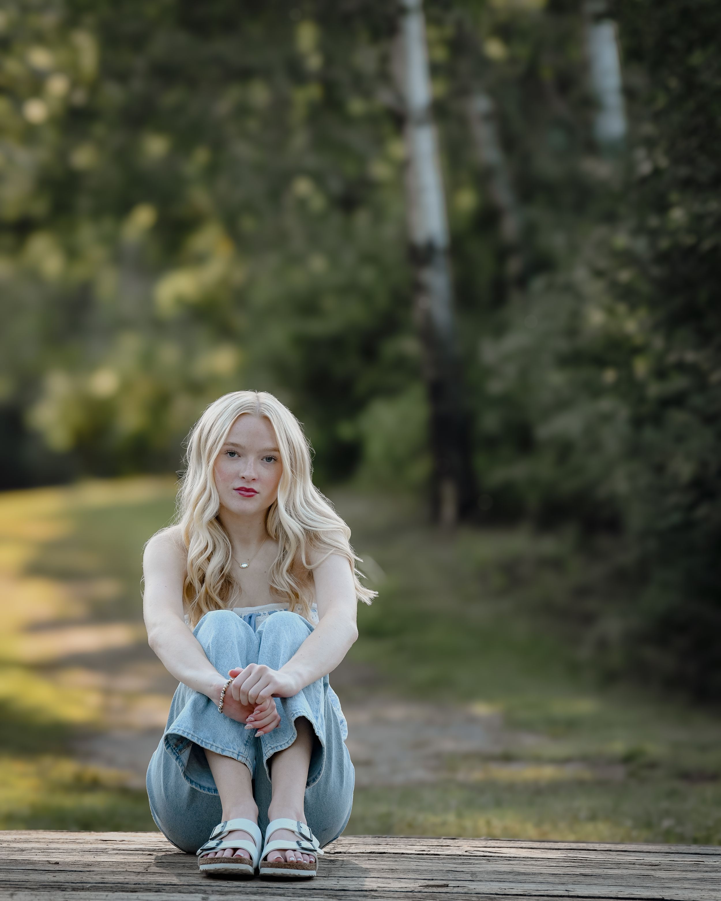Blonde woman in light blue outfit sitting on wooden path surrounded by green trees.