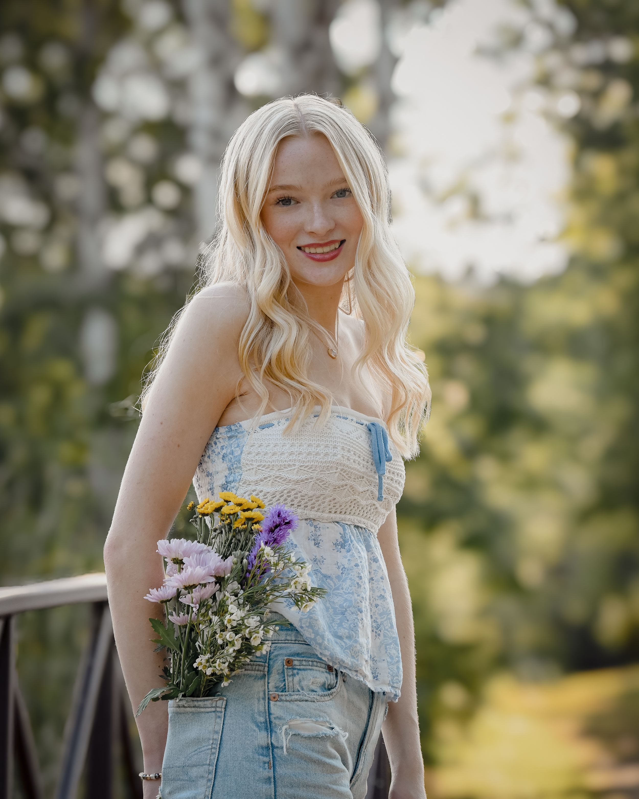 Smiling blonde woman holding wildflowers in denim overalls on a sunny outdoor bridge.