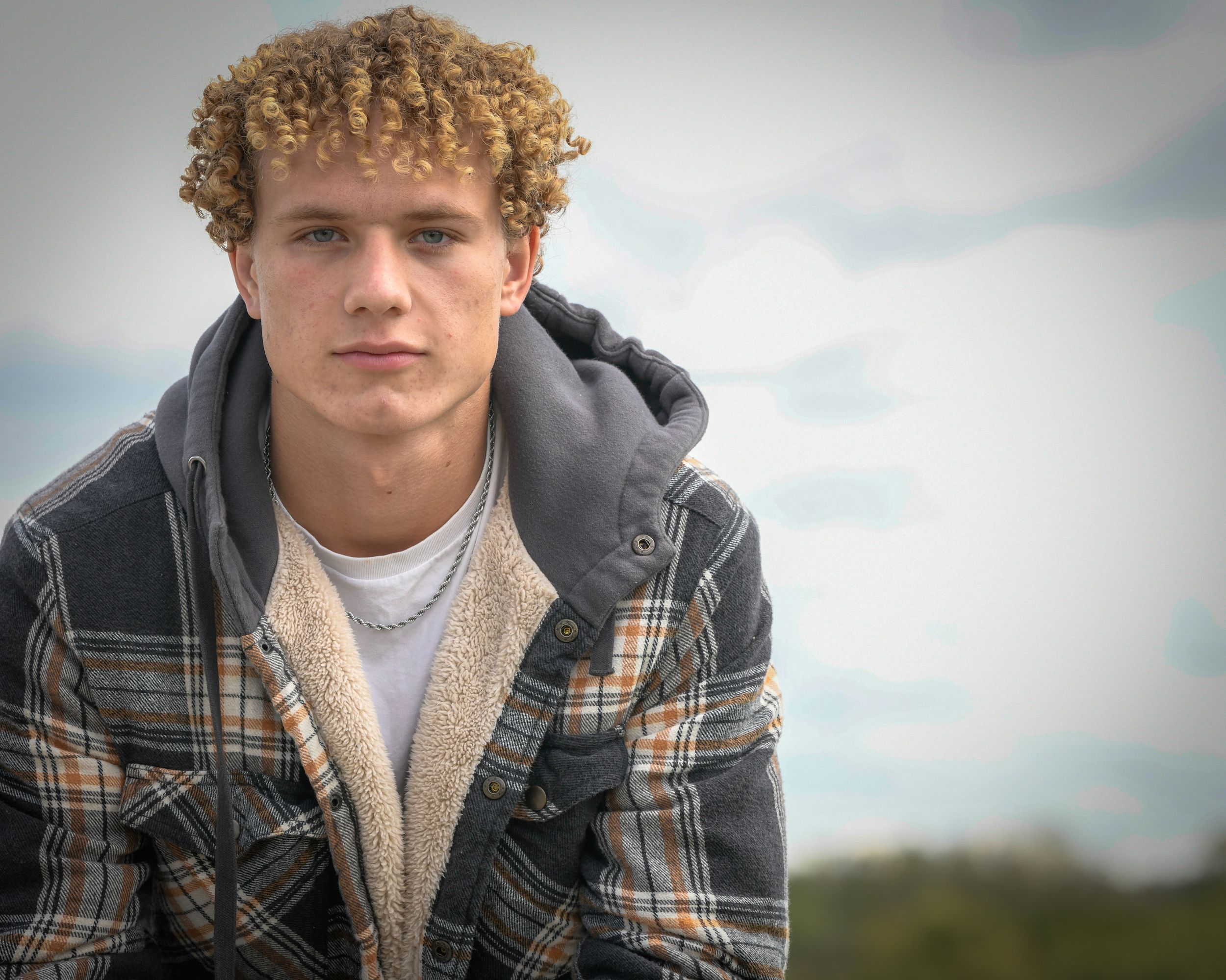 Young man with curly blonde hair wearing a plaid flannel hoodie jacket outdoors with cloudy sky background.