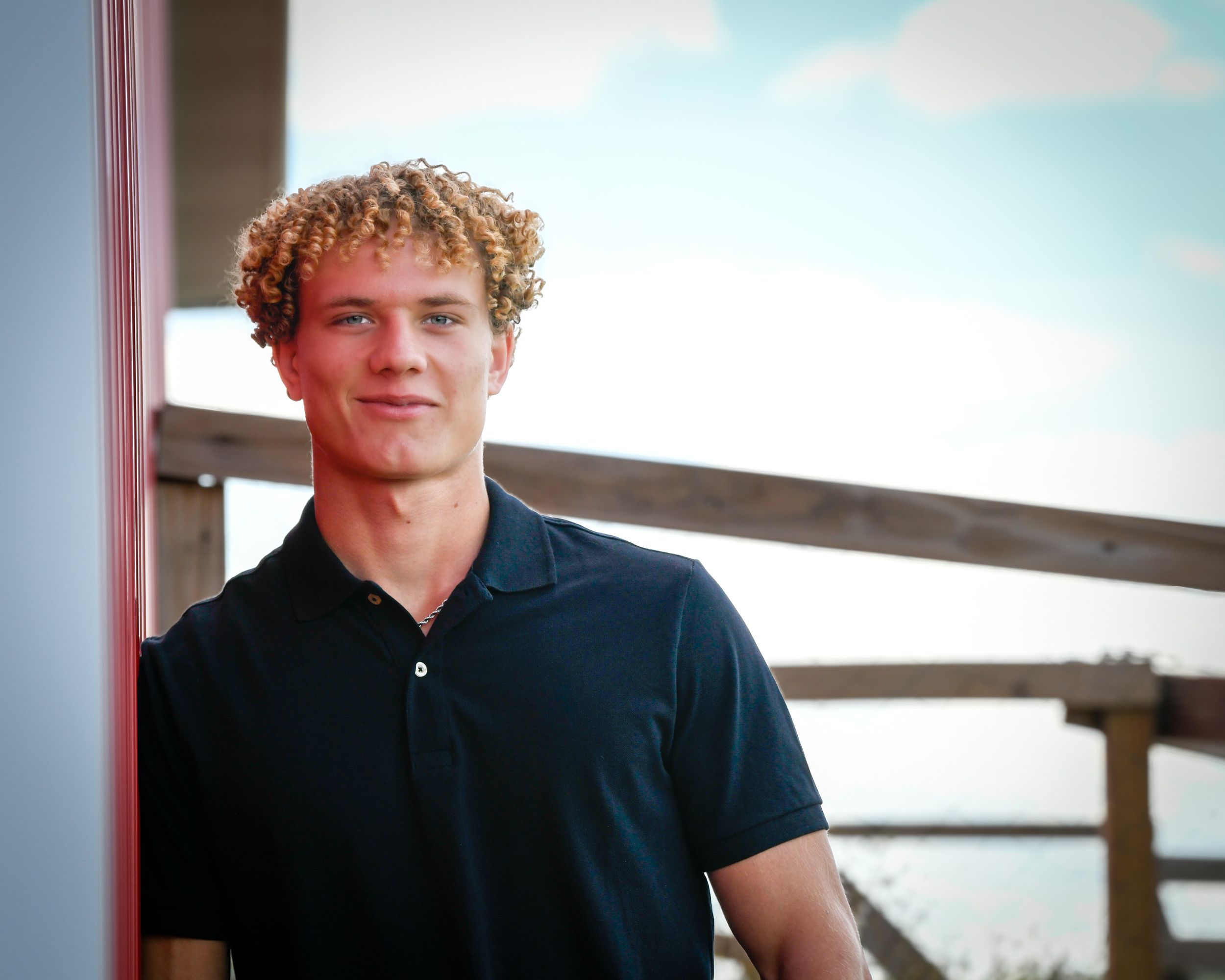 Young man with curly blonde hair wearing a black polo shirt, smiling outdoors near a wooden railing.
