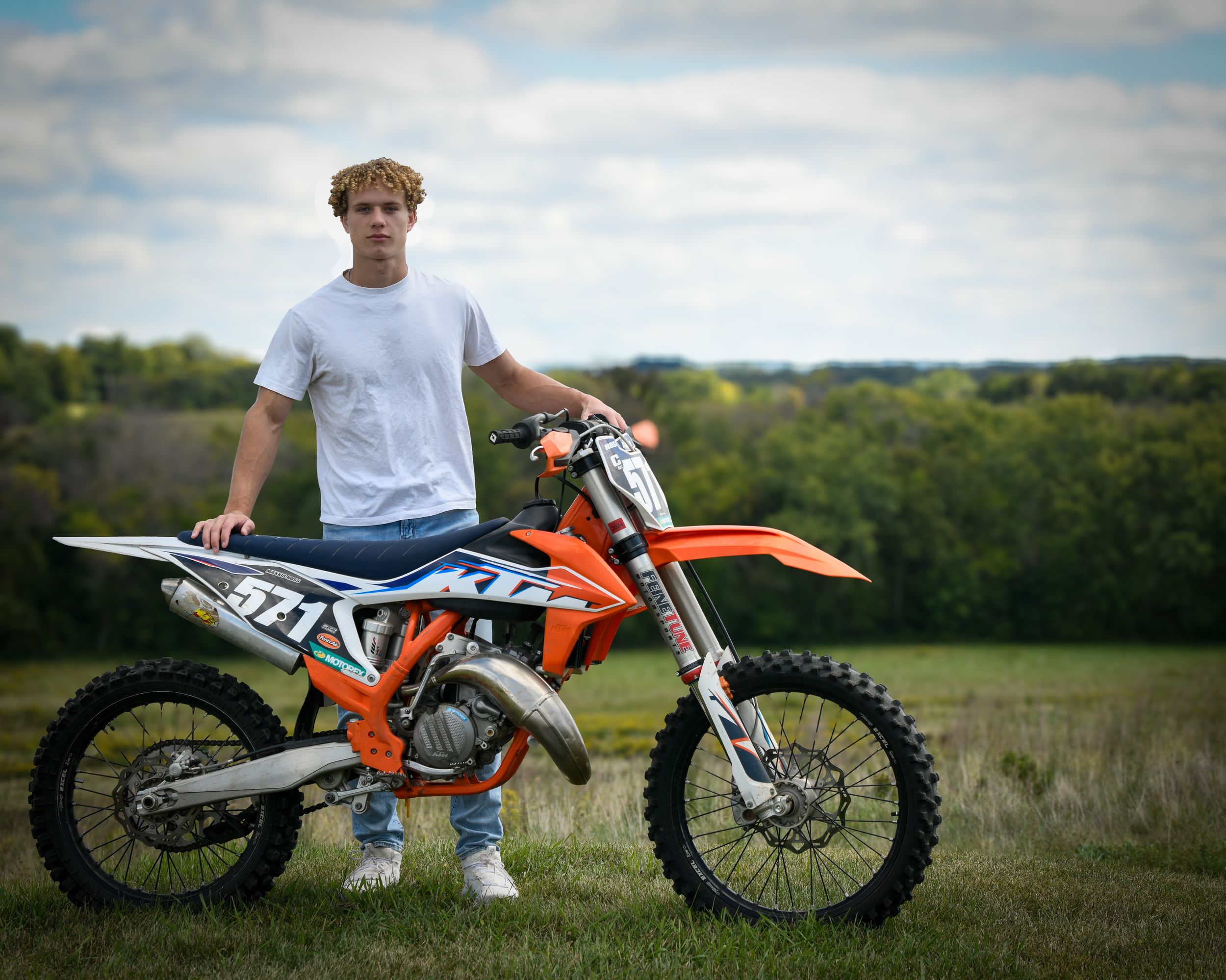 Young man poses with orange KTM dirt bike in a green field with trees and cloudy sky backdrop.
