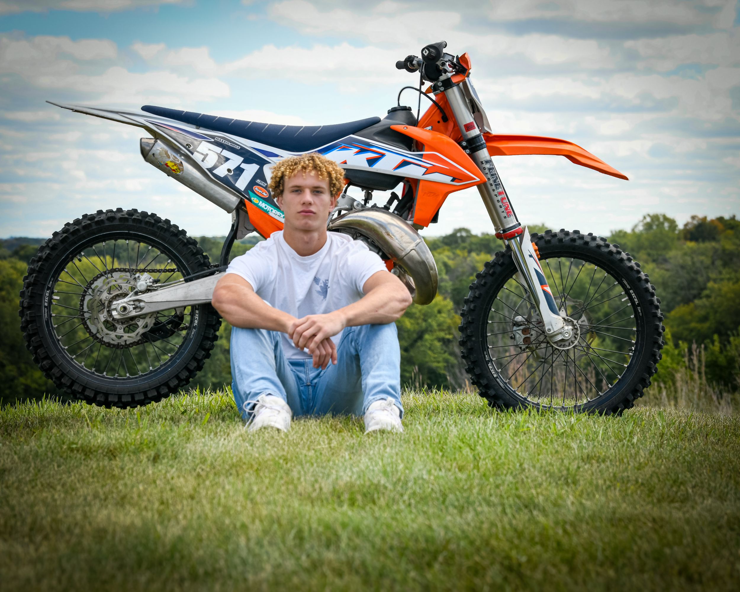 Teen boy sitting in grass in front of orange KTM dirt bike on a cloudy day outdoors.