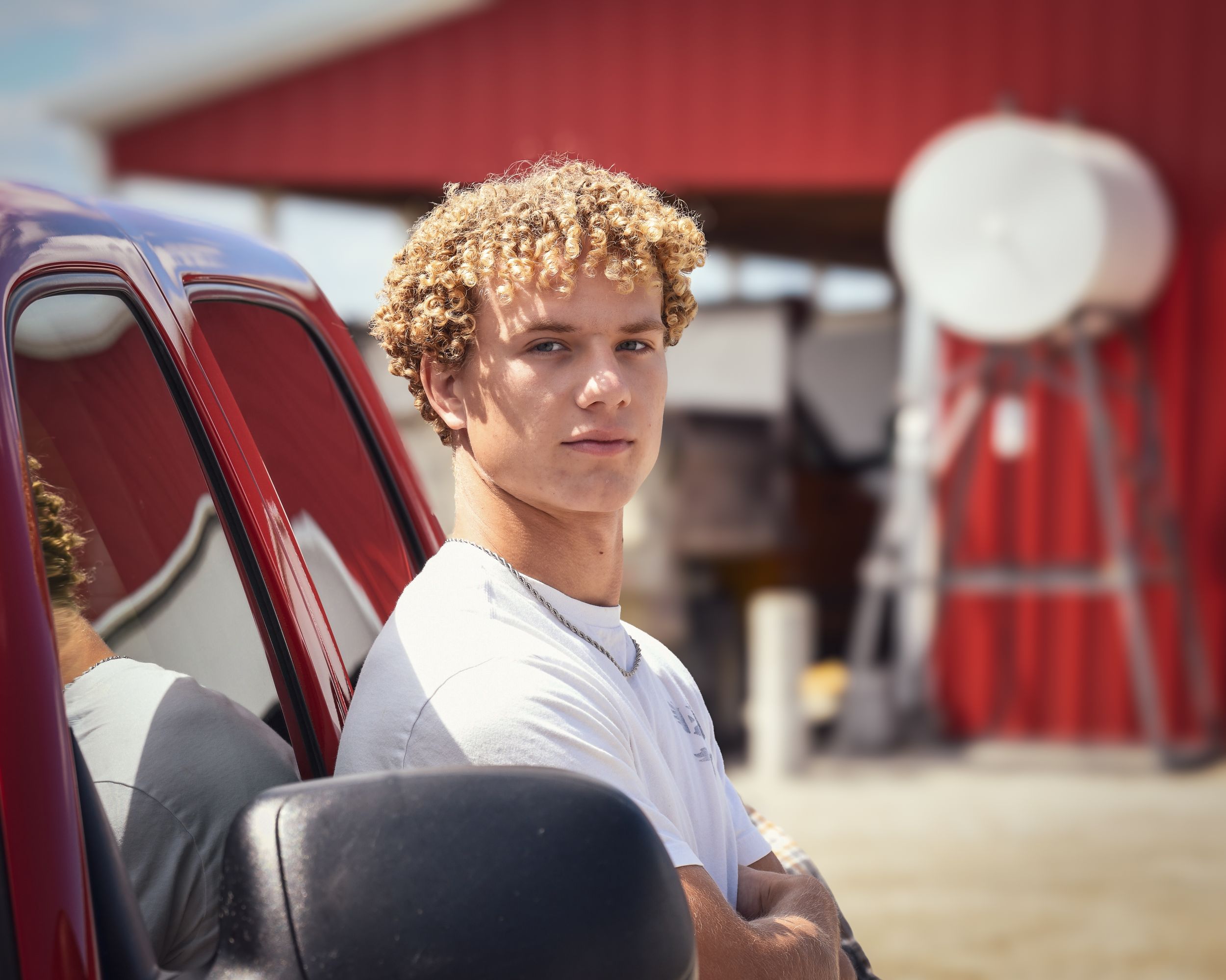 Young man with curly blonde hair stands by a red truck on a farm, arms crossed, wearing a white t-shirt.
