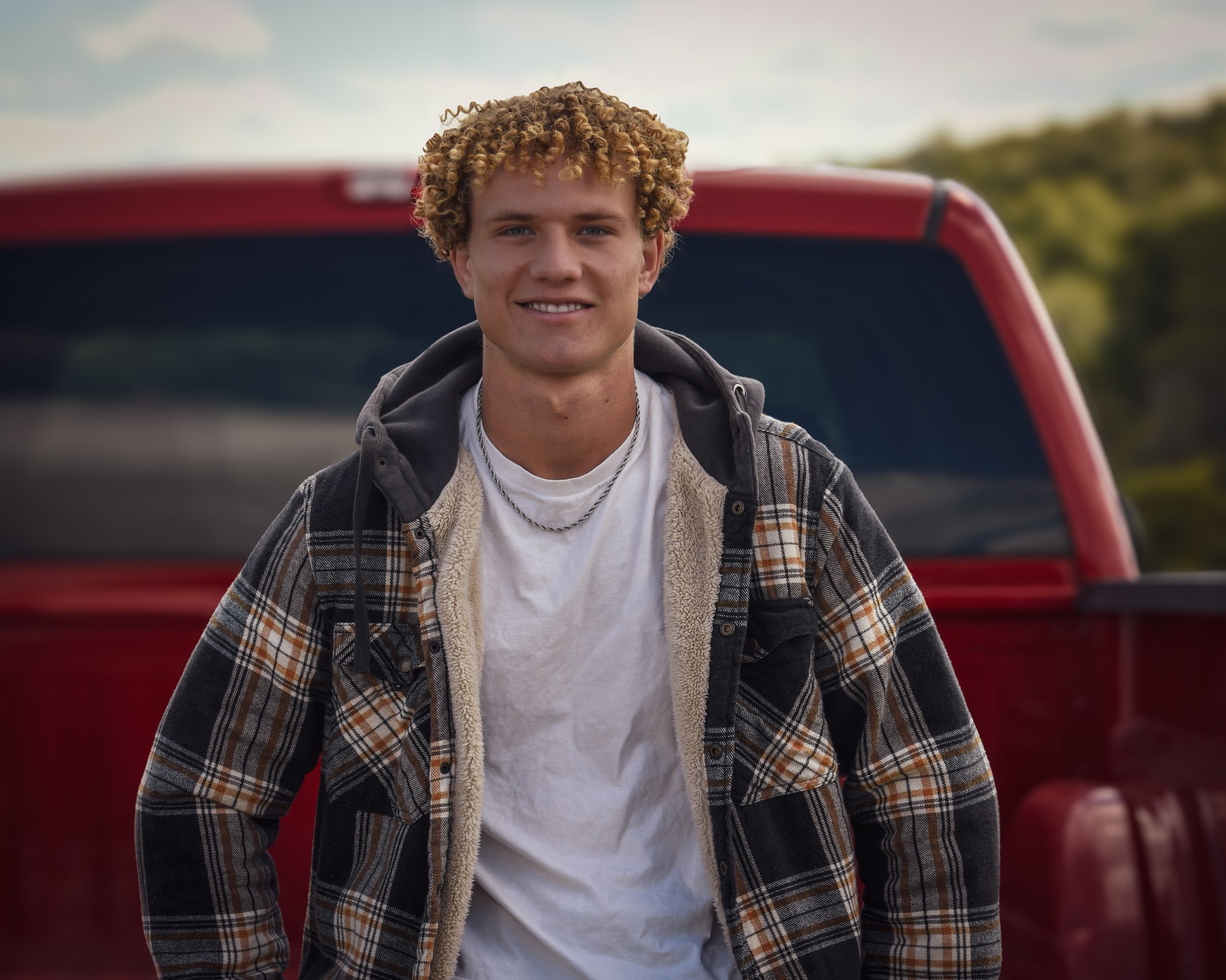 Smiling young man with curly hair in plaid flannel jacket leaning against a red pickup truck outdoors.