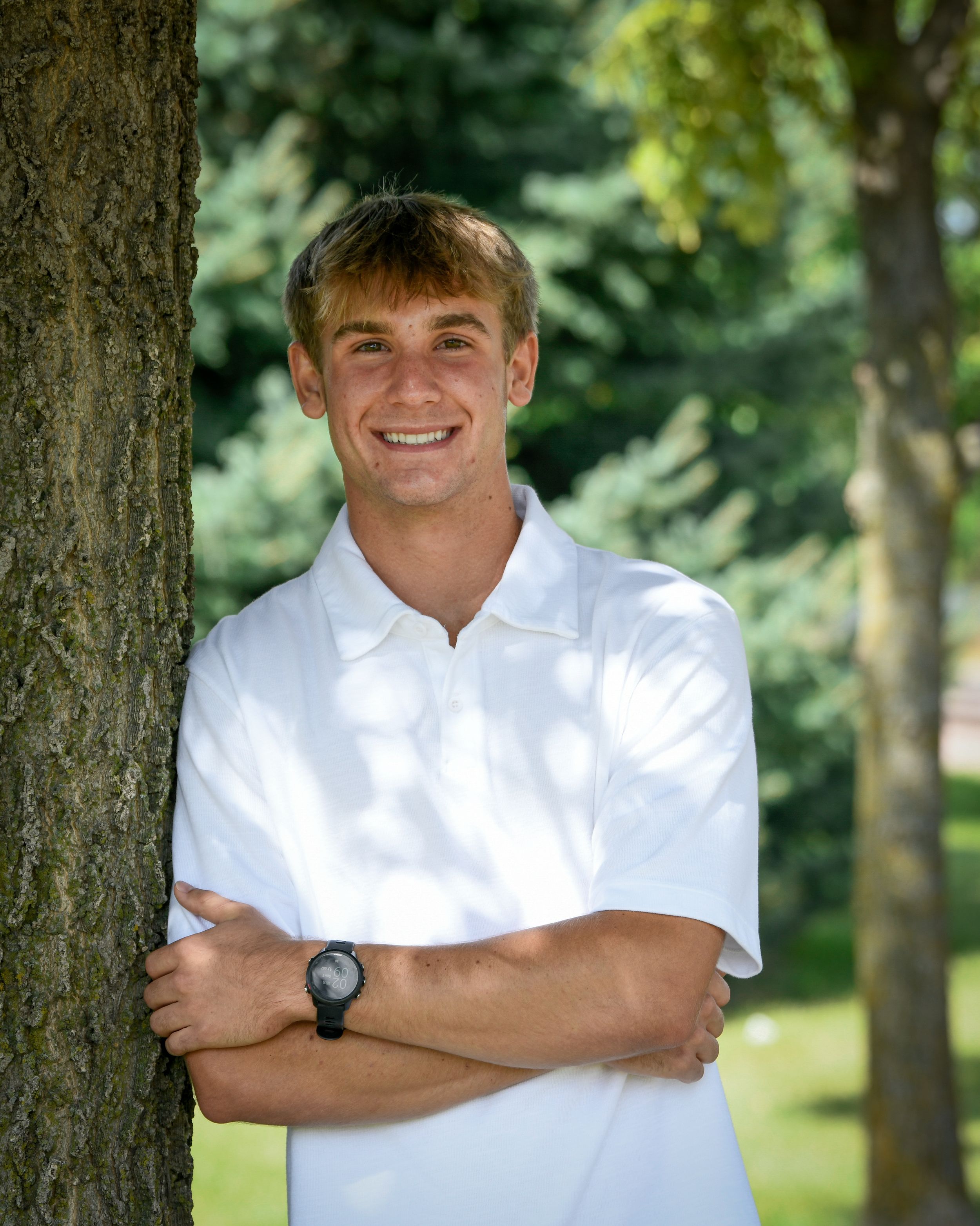 Smiling young man in white polo shirt leaning against tree with arms crossed in outdoor park setting.