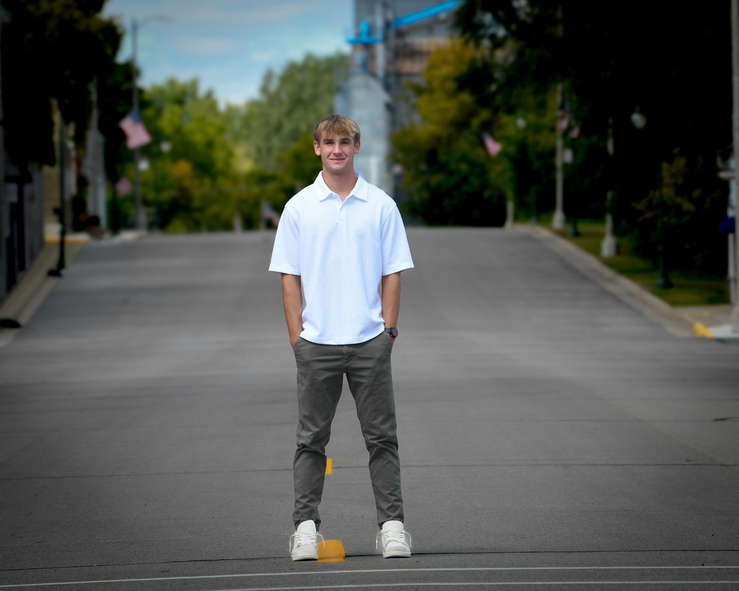 Young man in white polo shirt and gray pants standing confidently on an empty street lined with trees.