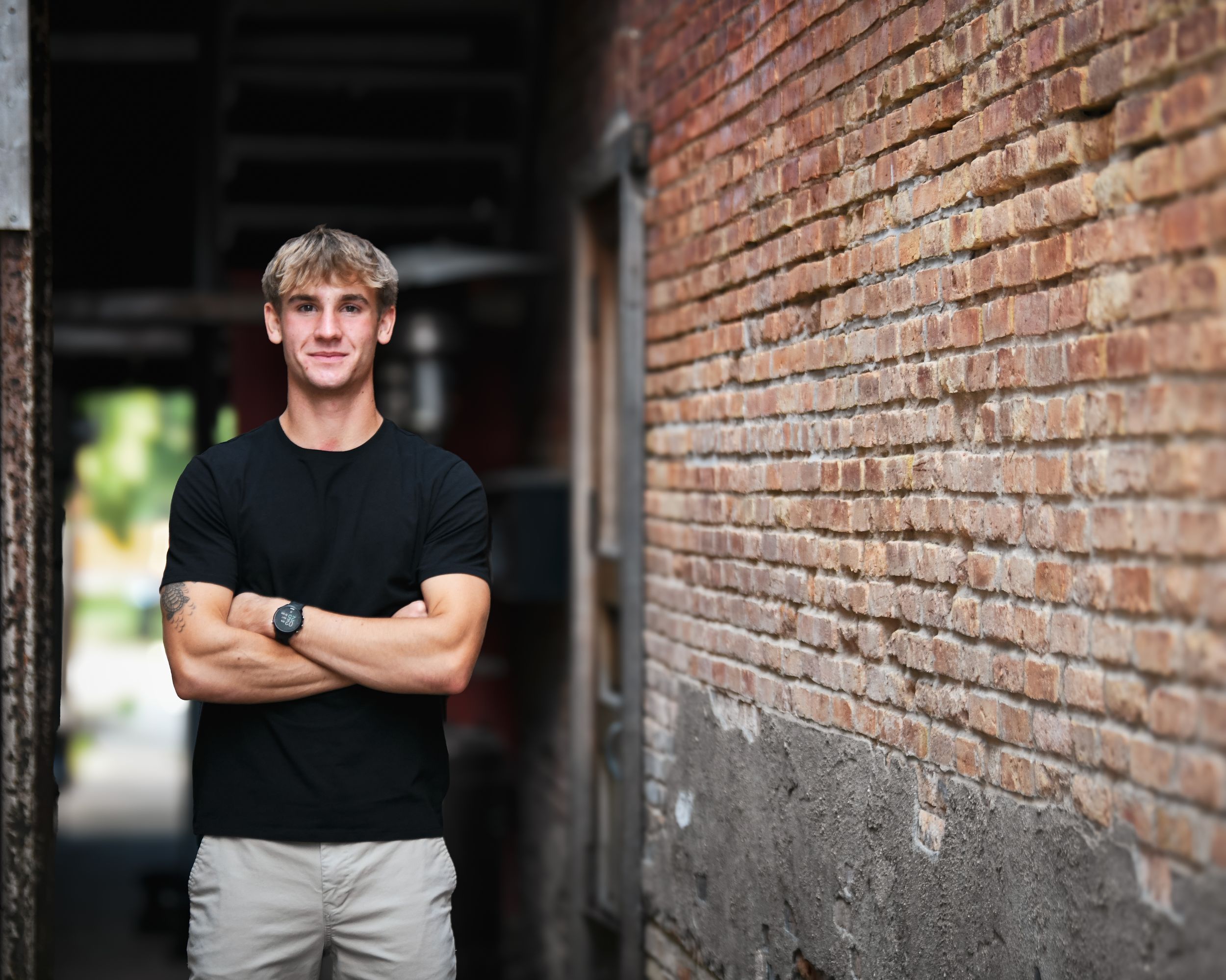 Young man in black t-shirt with arms crossed leaning against a rustic brick wall in an urban alley.