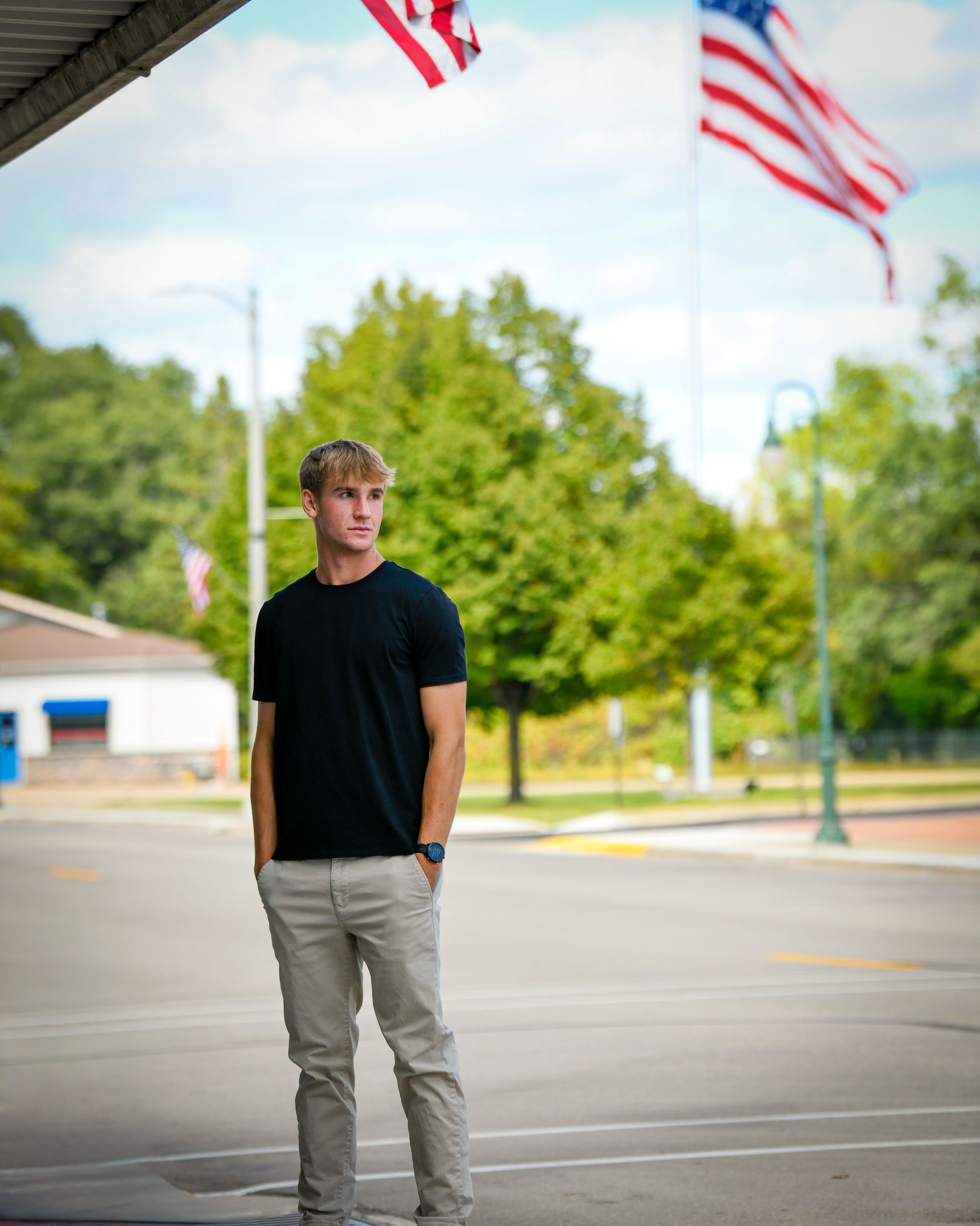 Young man in black t-shirt and khaki pants standing outdoors near American flags on a cloudy day.