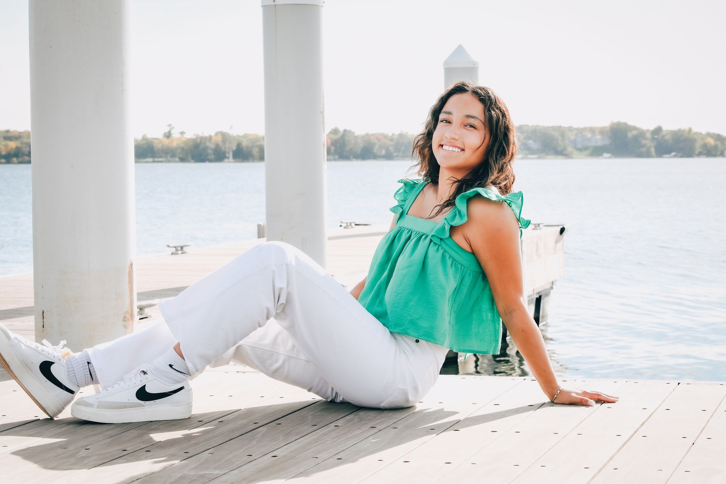 Woman in green top and white jeans smiling on a sunny waterfront dock, wearing Nike sneakers.