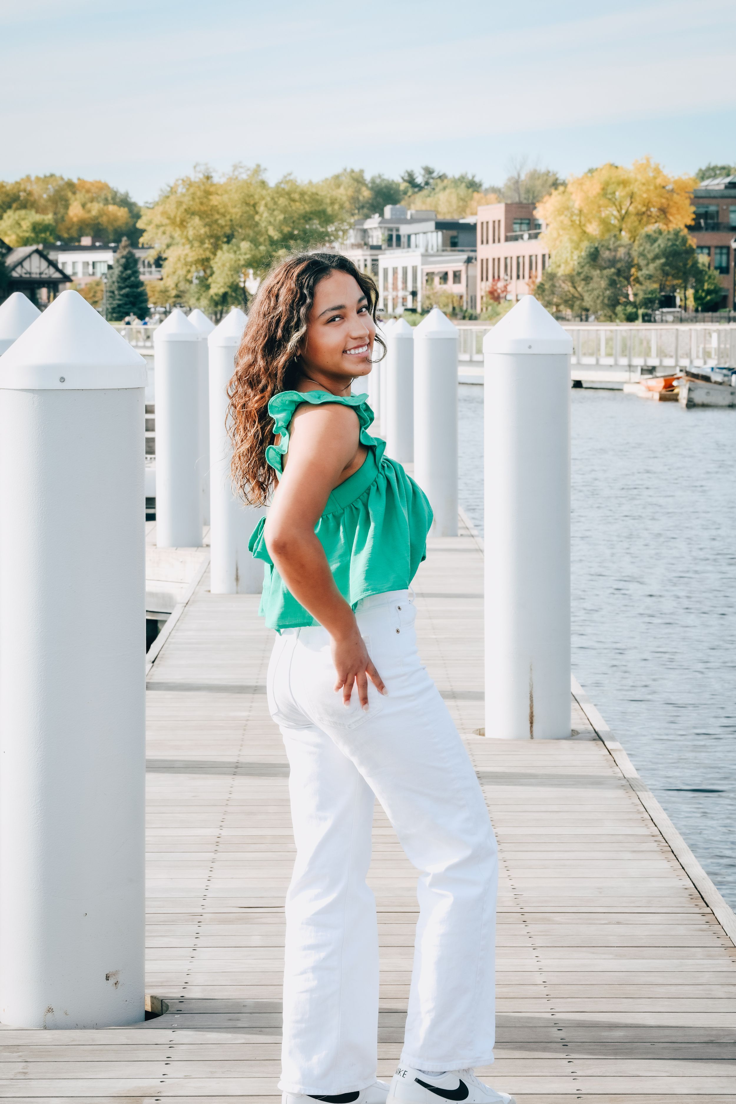 Young woman in green top and white pants smiling on a marina dock with water and buildings in background.