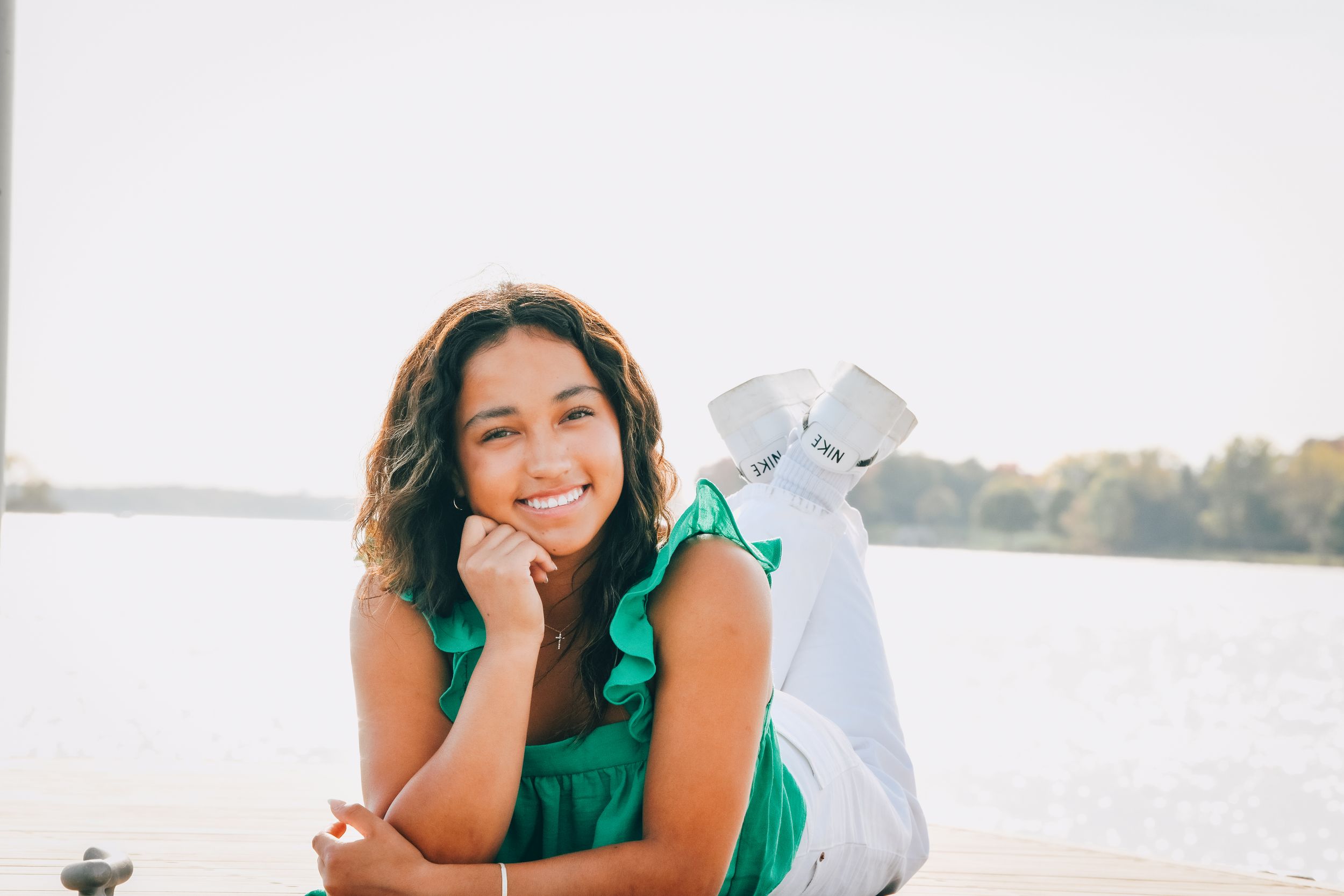 Smiling young woman in green top and white jeans posing on a dock by a lake on a sunny day.