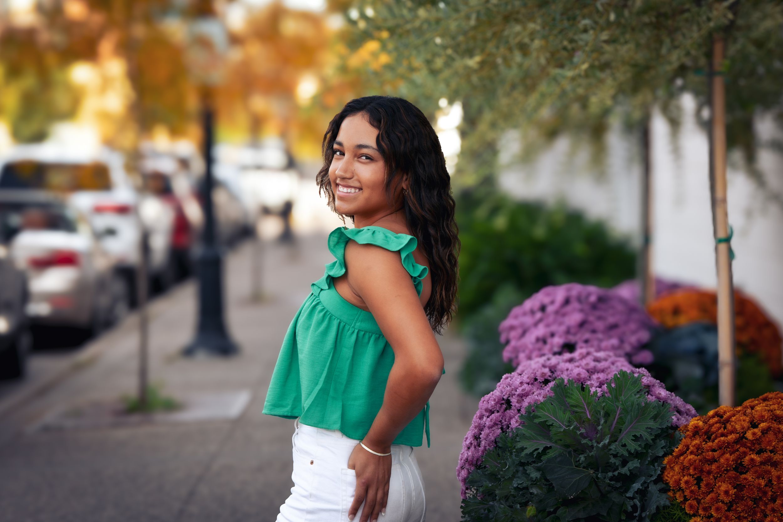 Young woman in green ruffled top and white pants smiling on a city sidewalk with colorful mums nearby.