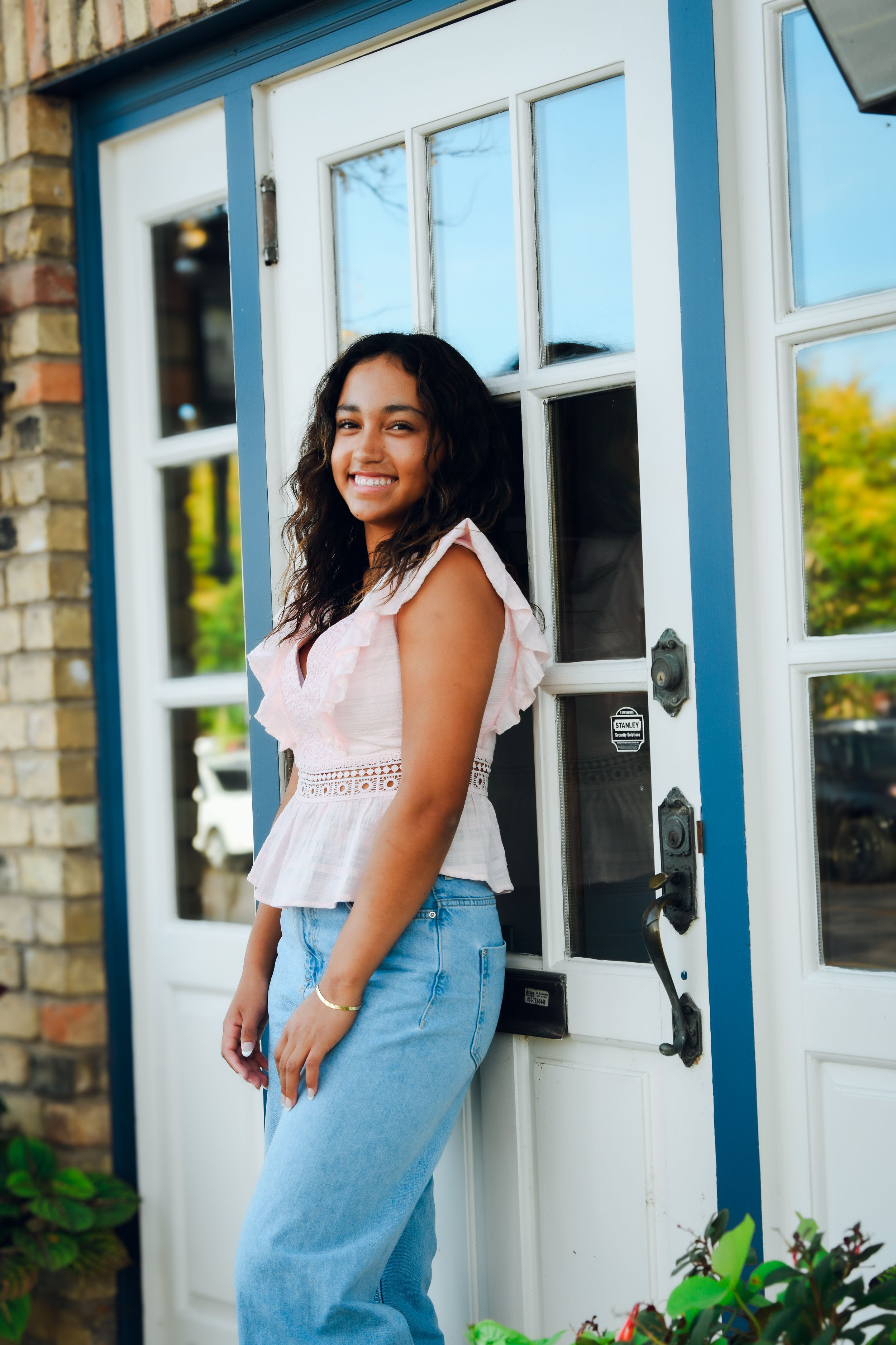 Young woman in pink top and jeans smiling by a blue and white door outdoors.