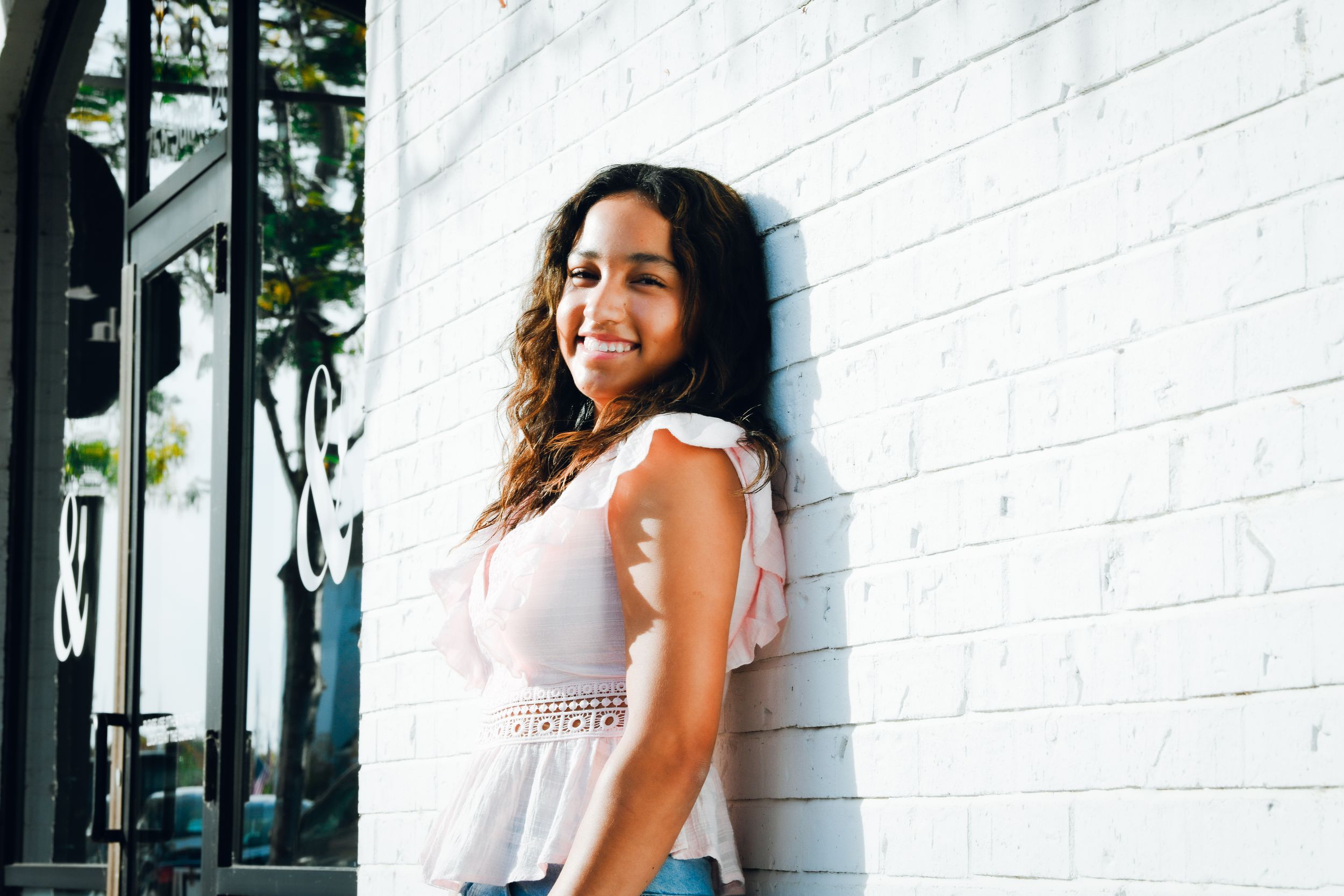Young woman with curly hair smiling confidently while leaning against a white brick wall outdoors.