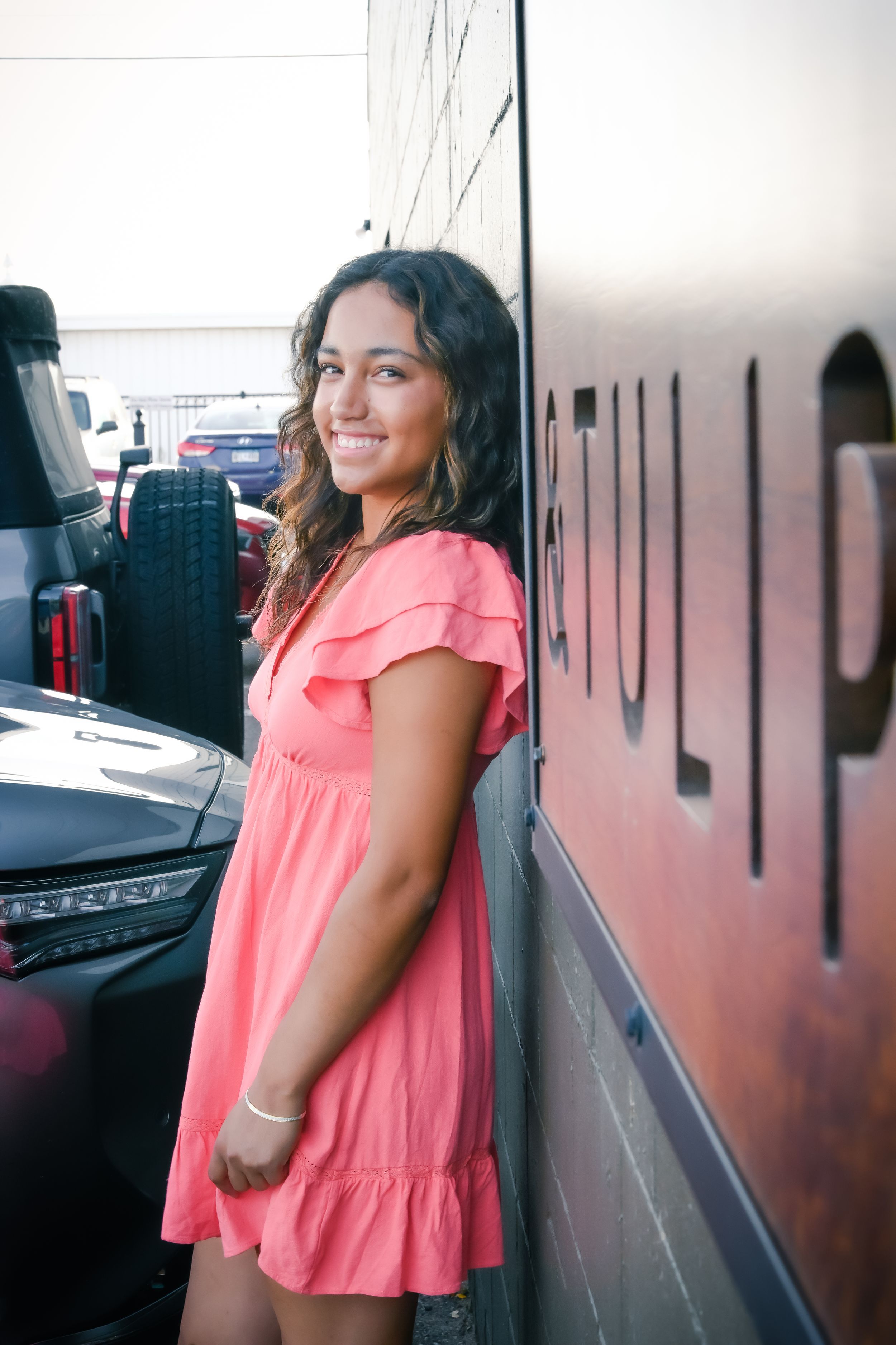 Young woman in pink ruffle dress smiling and leaning against a dark wall outdoors near parked vehicles.
