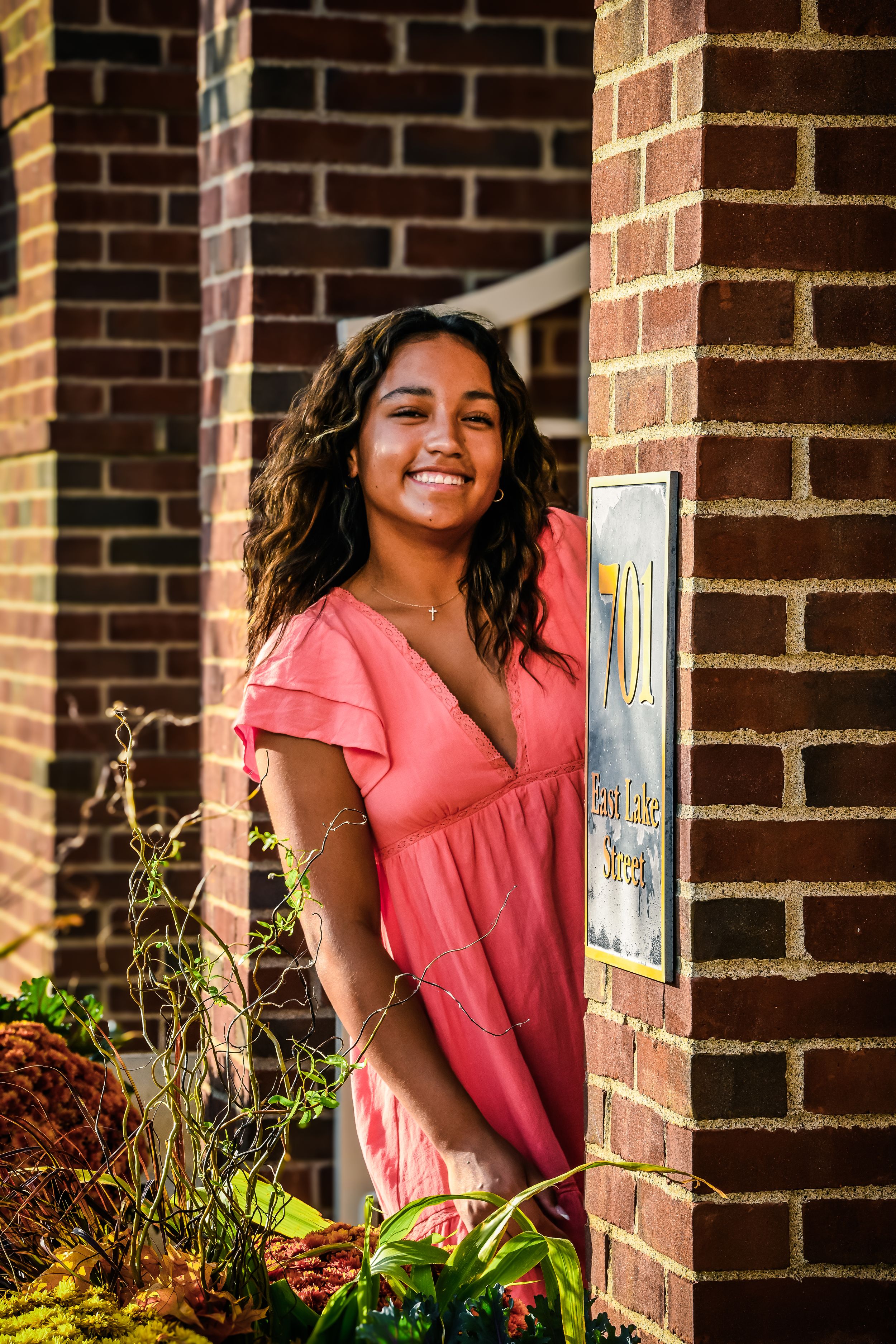 Smiling young woman in pink dress posing by brick building entrance with address number sign.