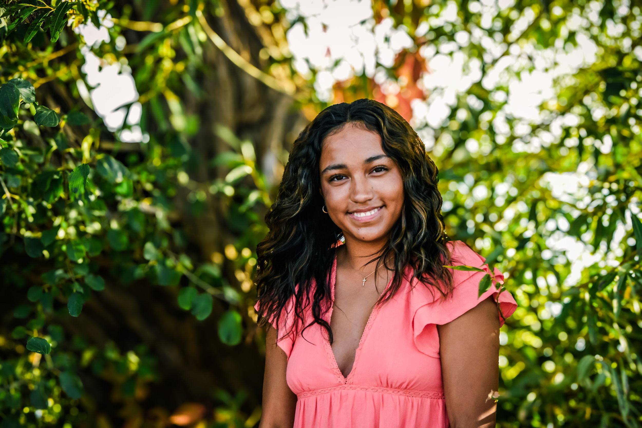 Smiling young woman in pink dress standing outdoors among lush green tropical trees in natural sunlight.