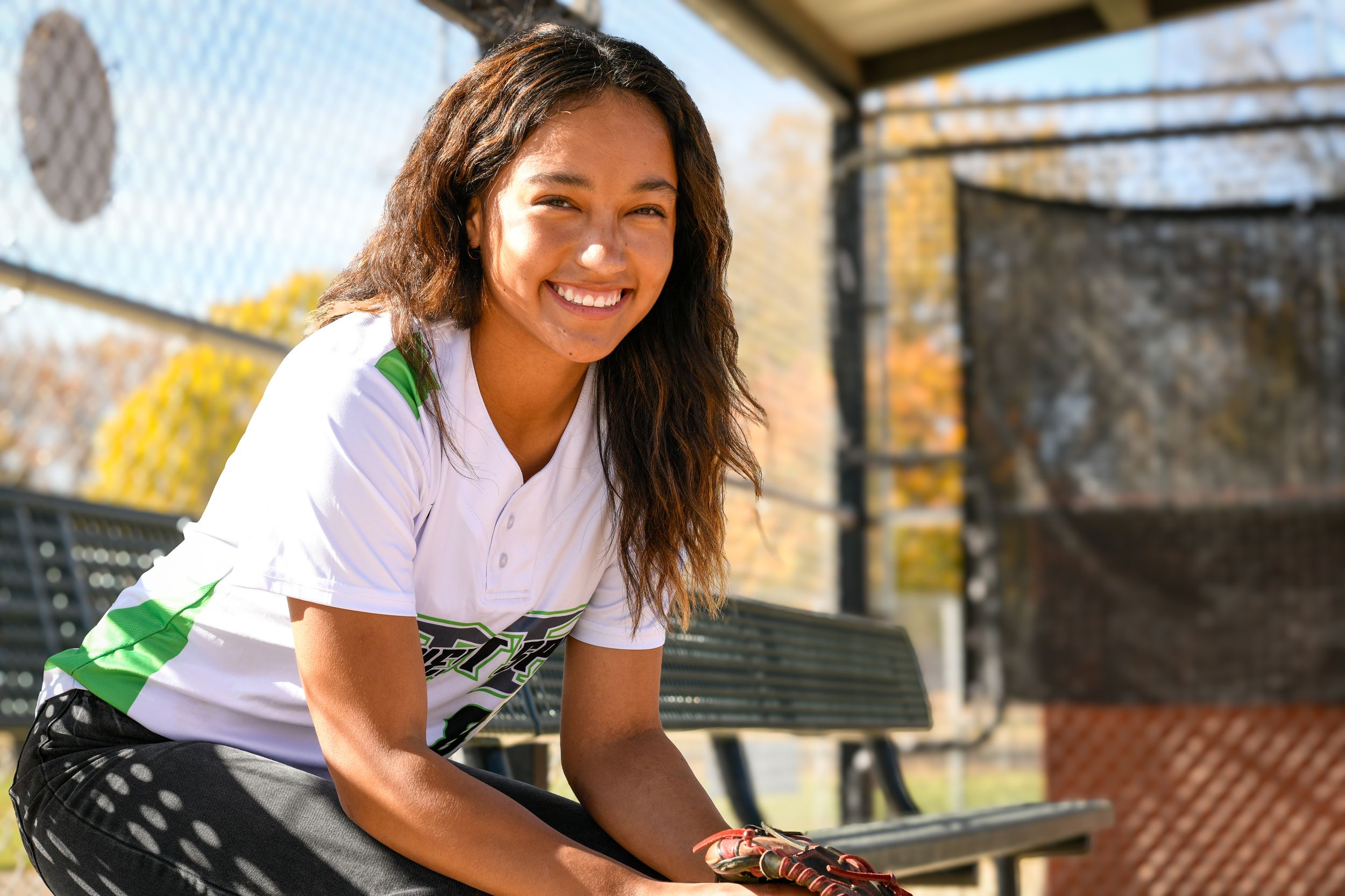 Smiling young female softball player in white jersey leaning on dugout fence on sunny autumn day.