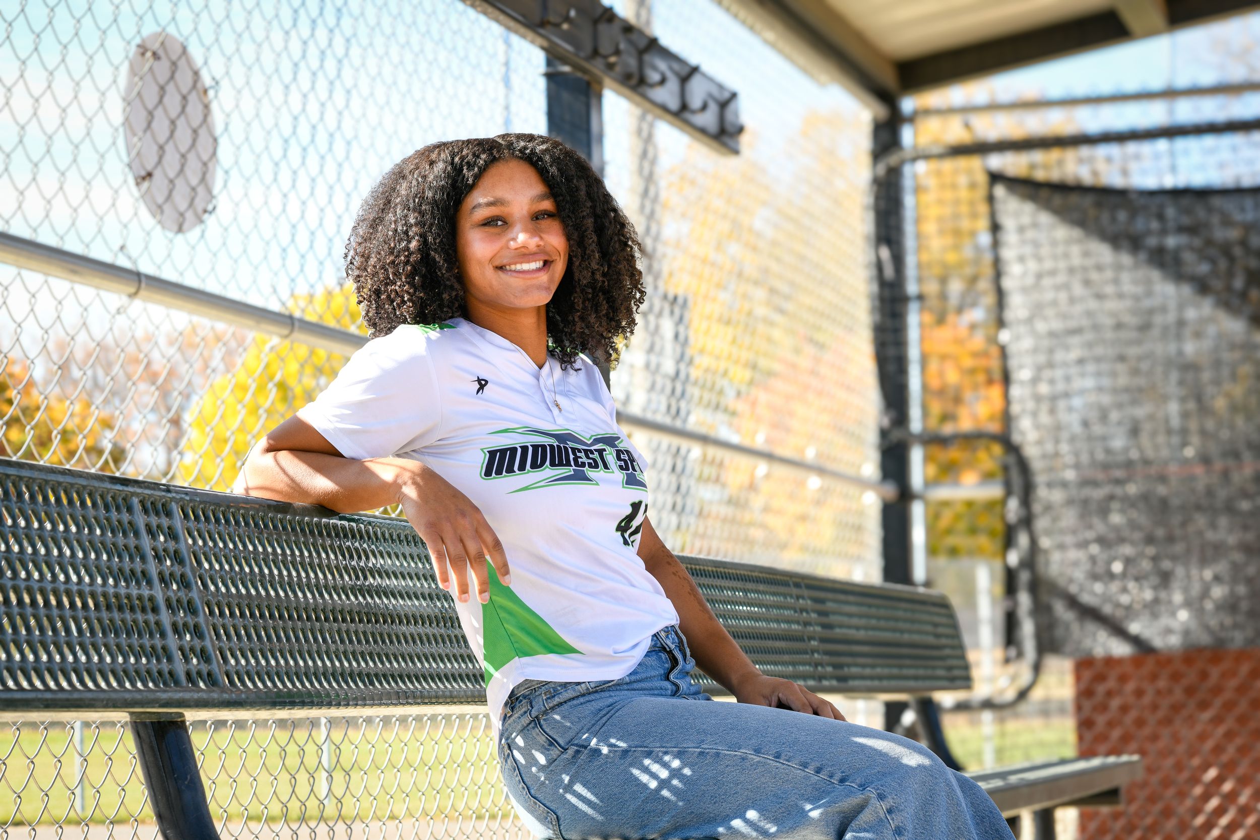 Young girl in Midwest softball jersey smiles while sitting on dugout bench at baseball field.