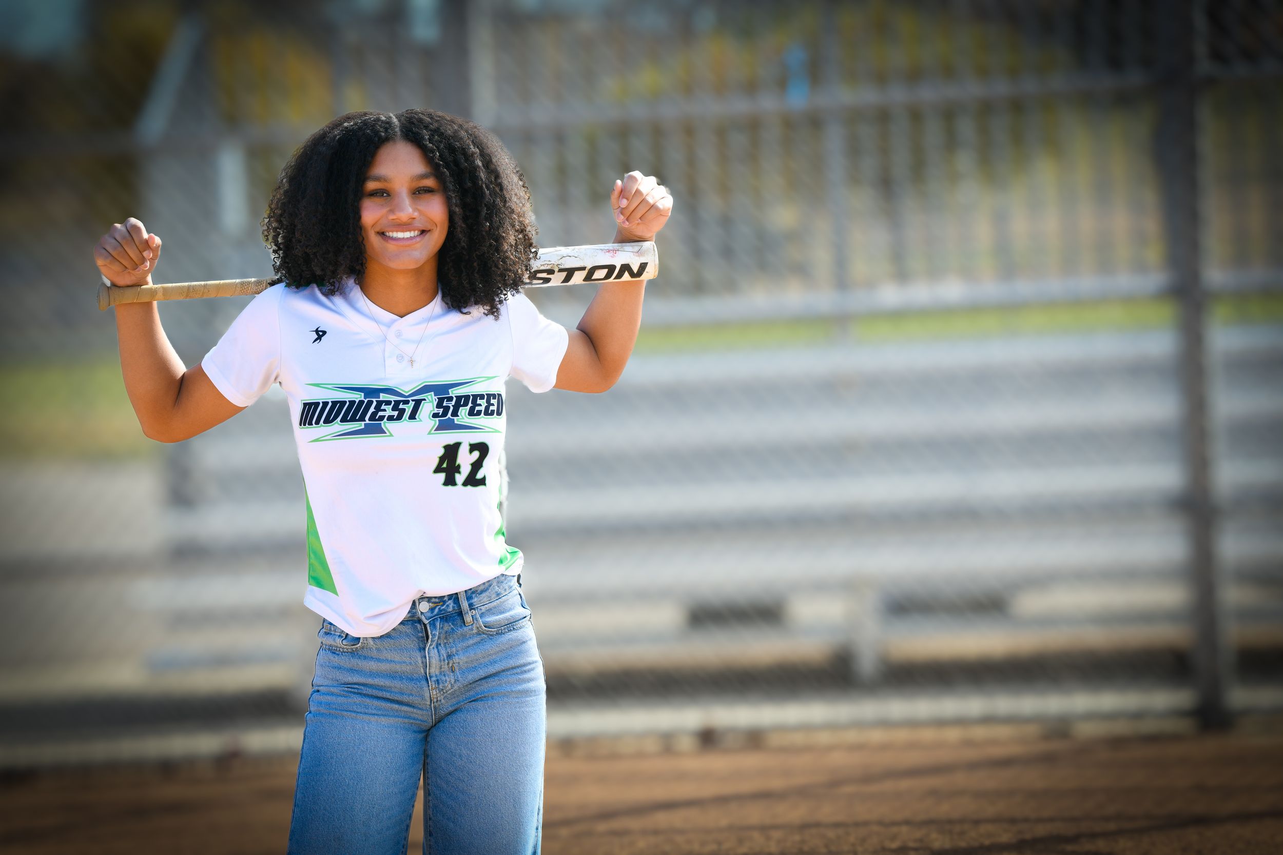 Young softball player in Midwest Speed #42 jersey poses with Easton bat on her shoulders at a baseball field.