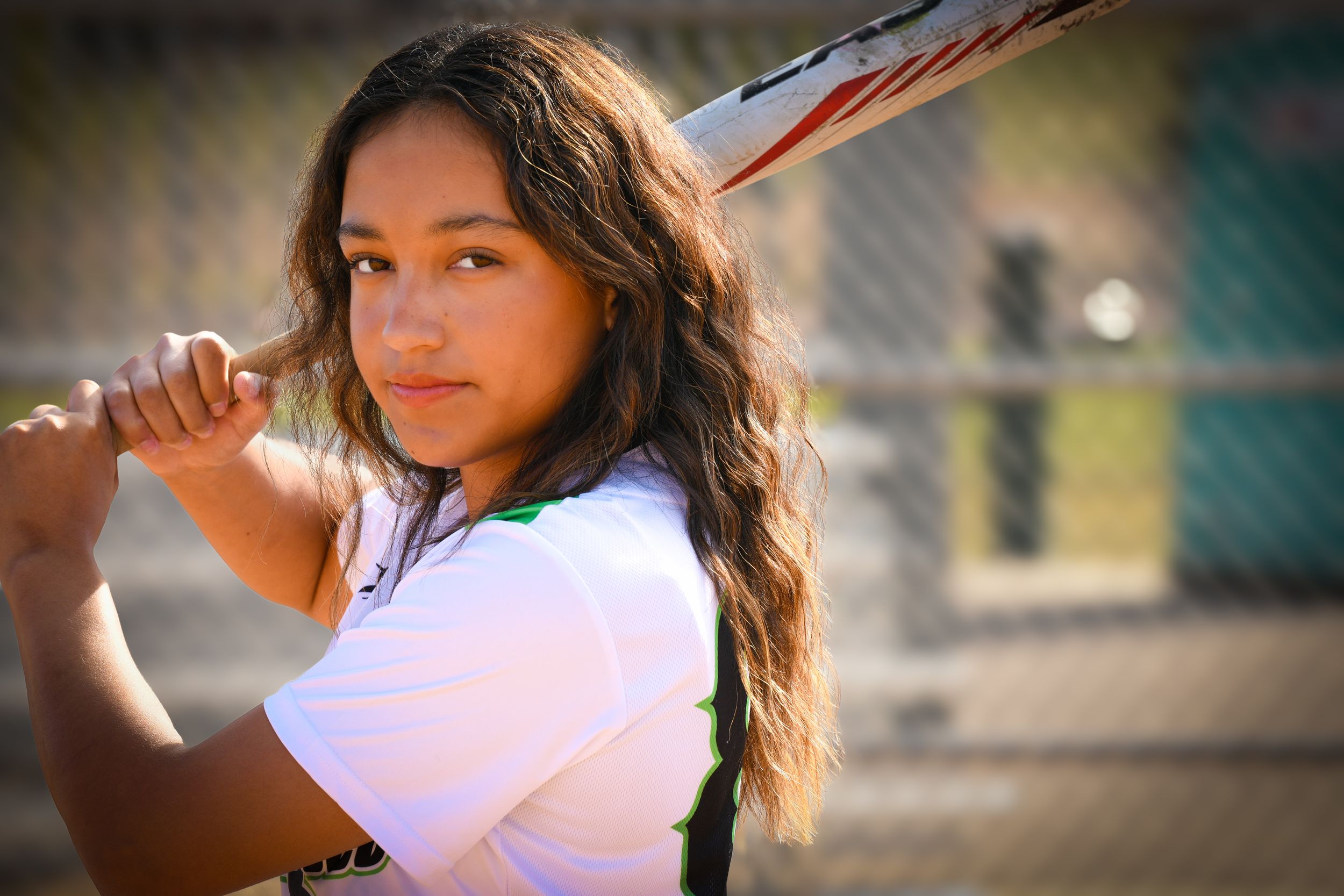 Young female softball player poses with bat over shoulder on outdoor field, wearing white jersey.