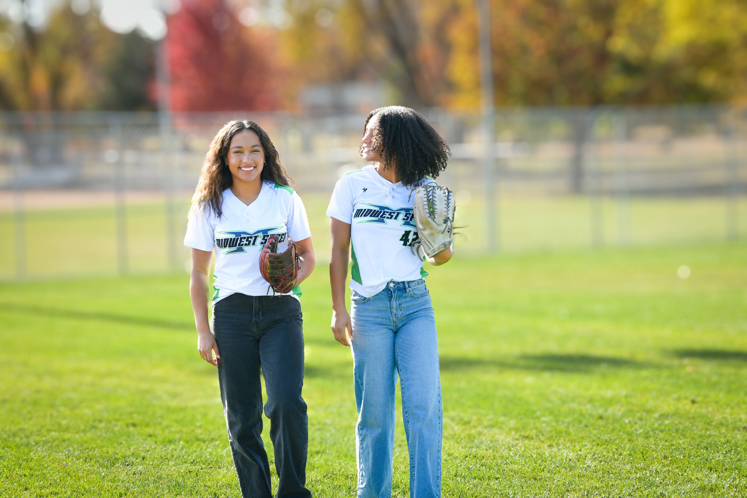 Two girls in white jerseys walk on a green field carrying softball gloves on a sunny autumn day.