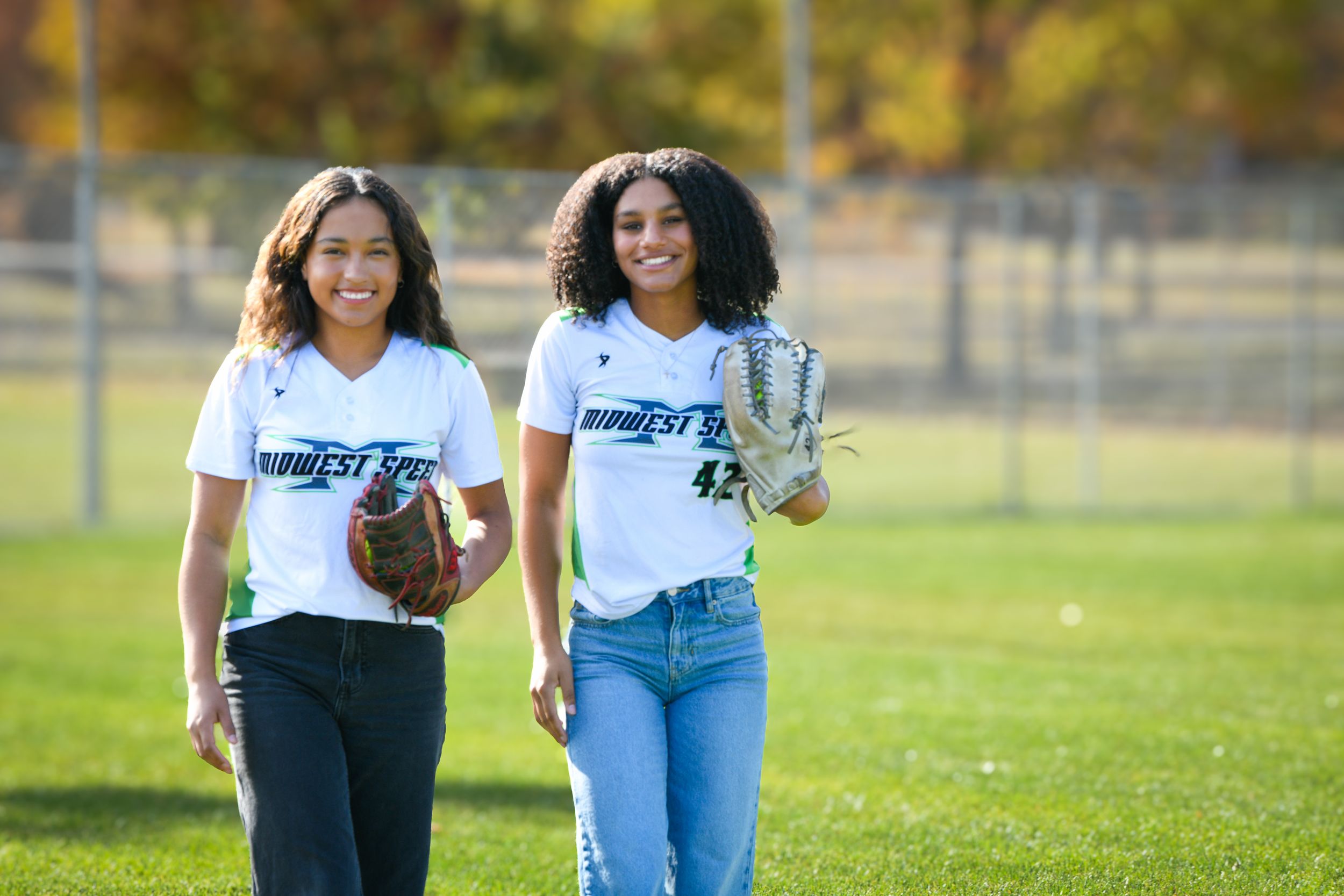 Two young women in softball uniforms holding gloves walk on a green field, smiling outdoors.