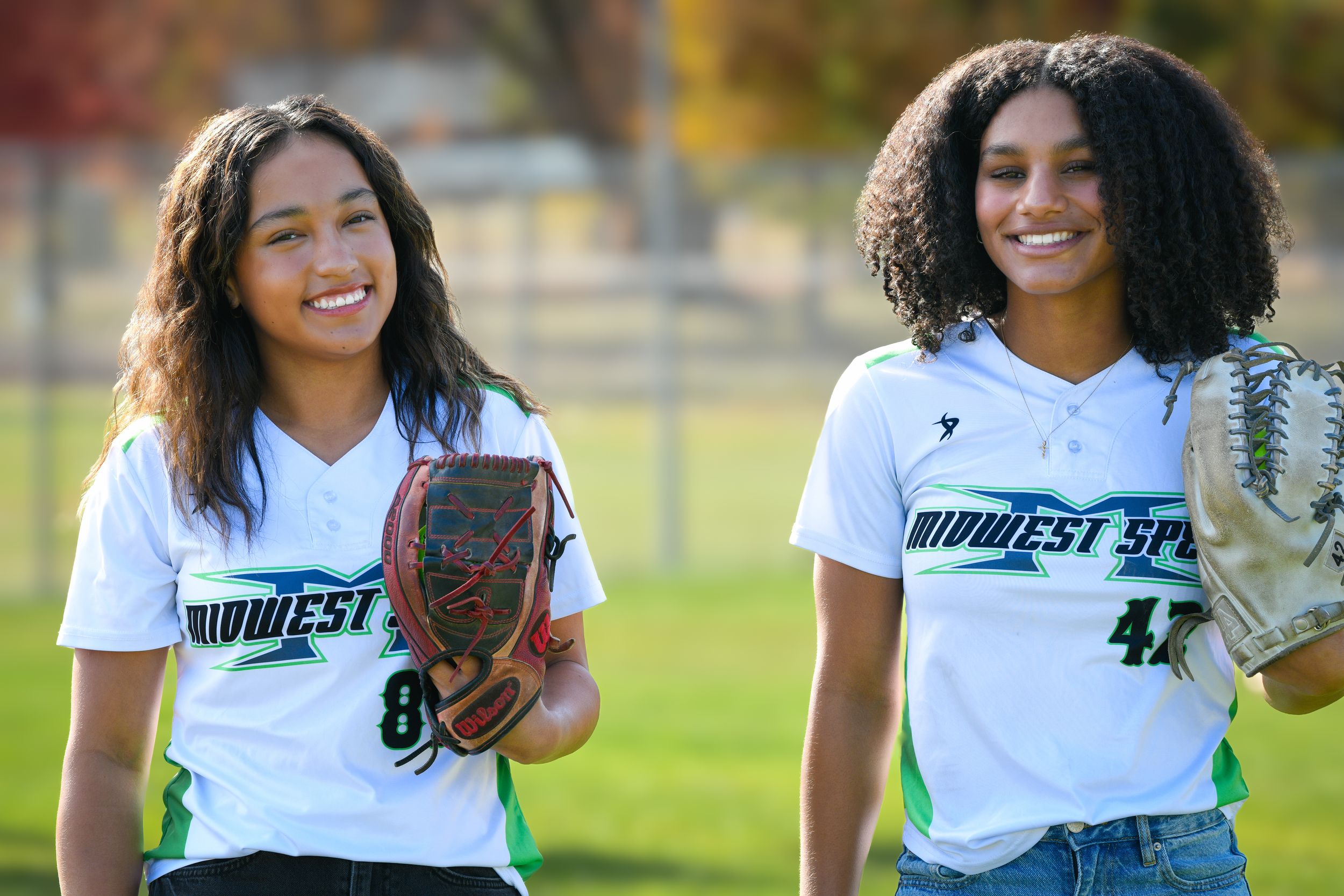 Two young softball players in Midwest Sports uniforms smiling on a field, one holding a glove.