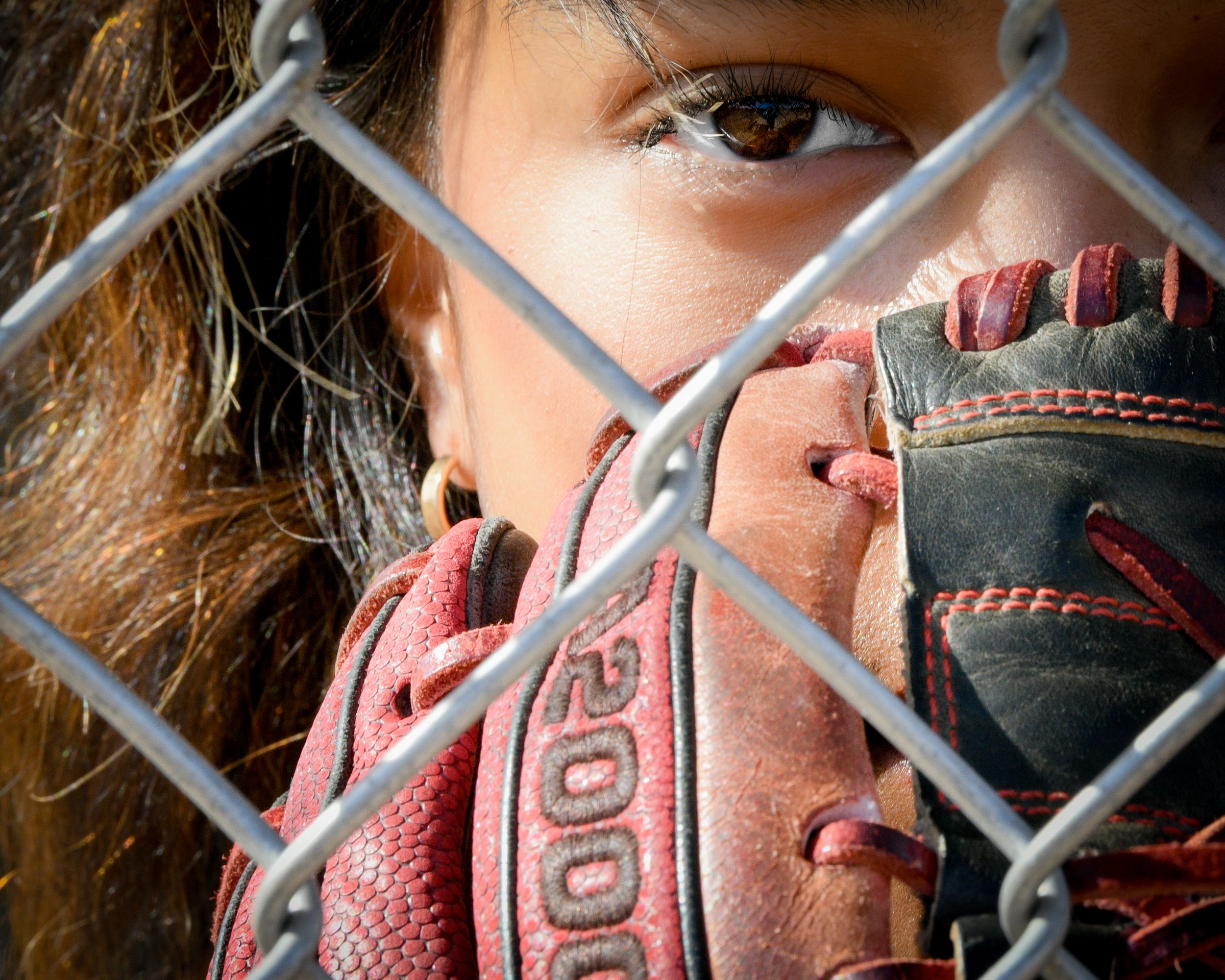 Young female softball player peers through chain-link fence holding a worn pink baseball glove up to her face.