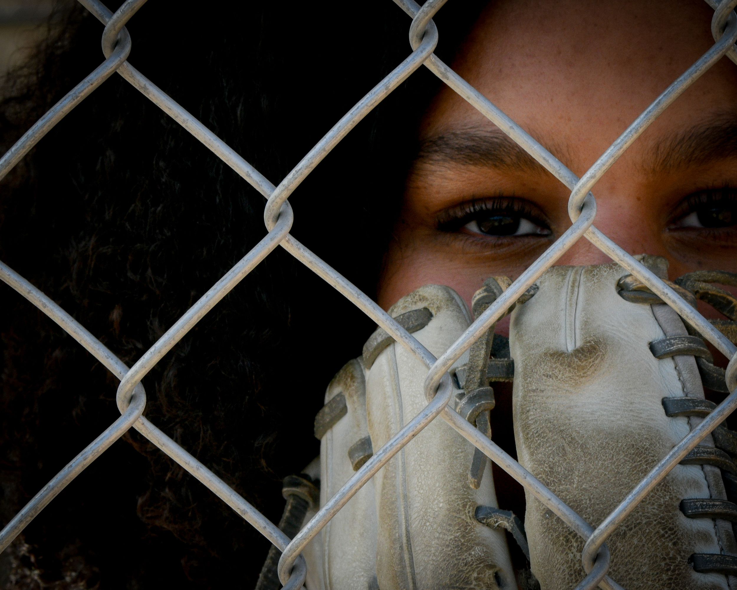 Young baseball player peering through chain-link fence holding a worn leather mitt up to their face.