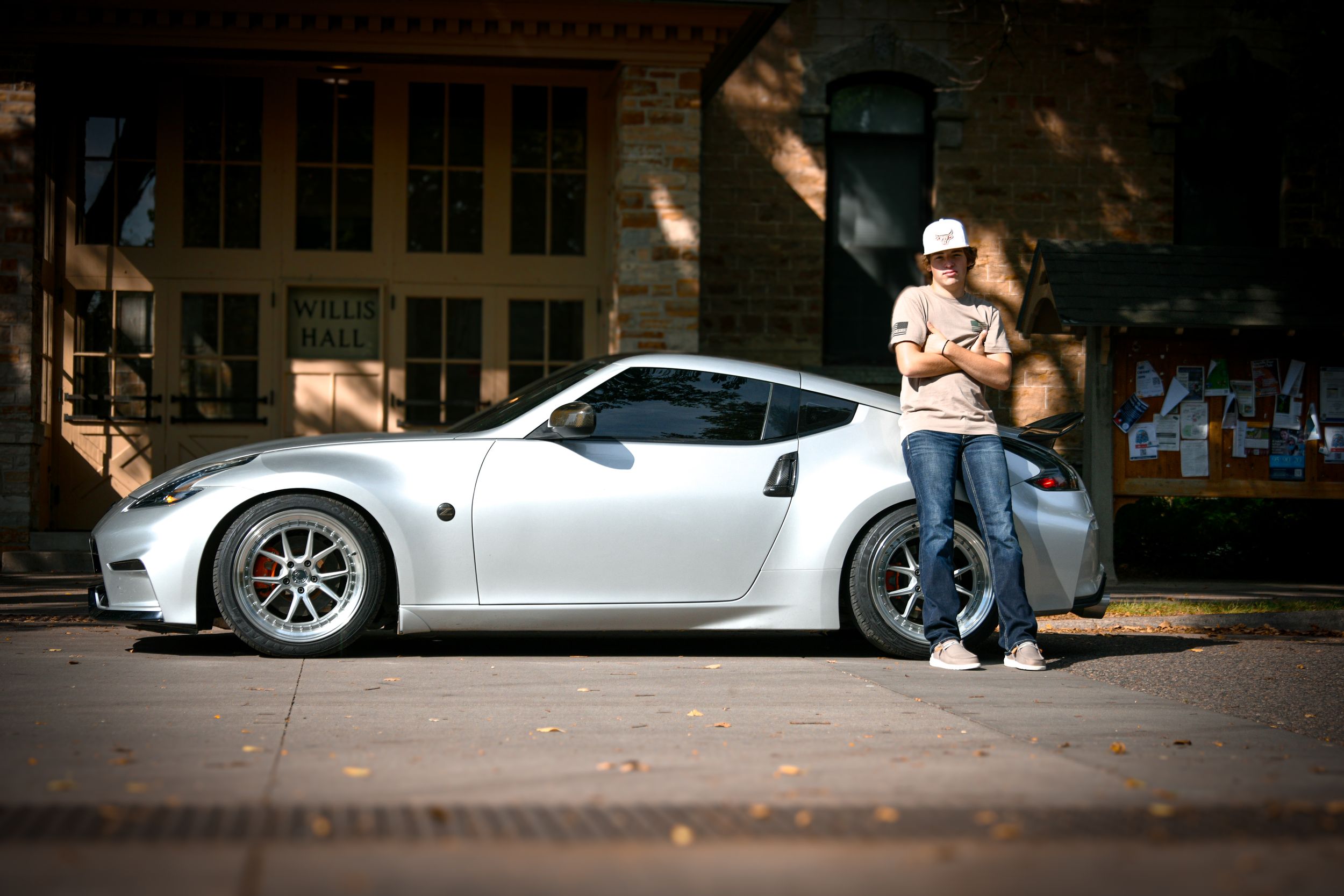 A man leans against a white Nissan 370Z sports car parked on a brick street near a building.