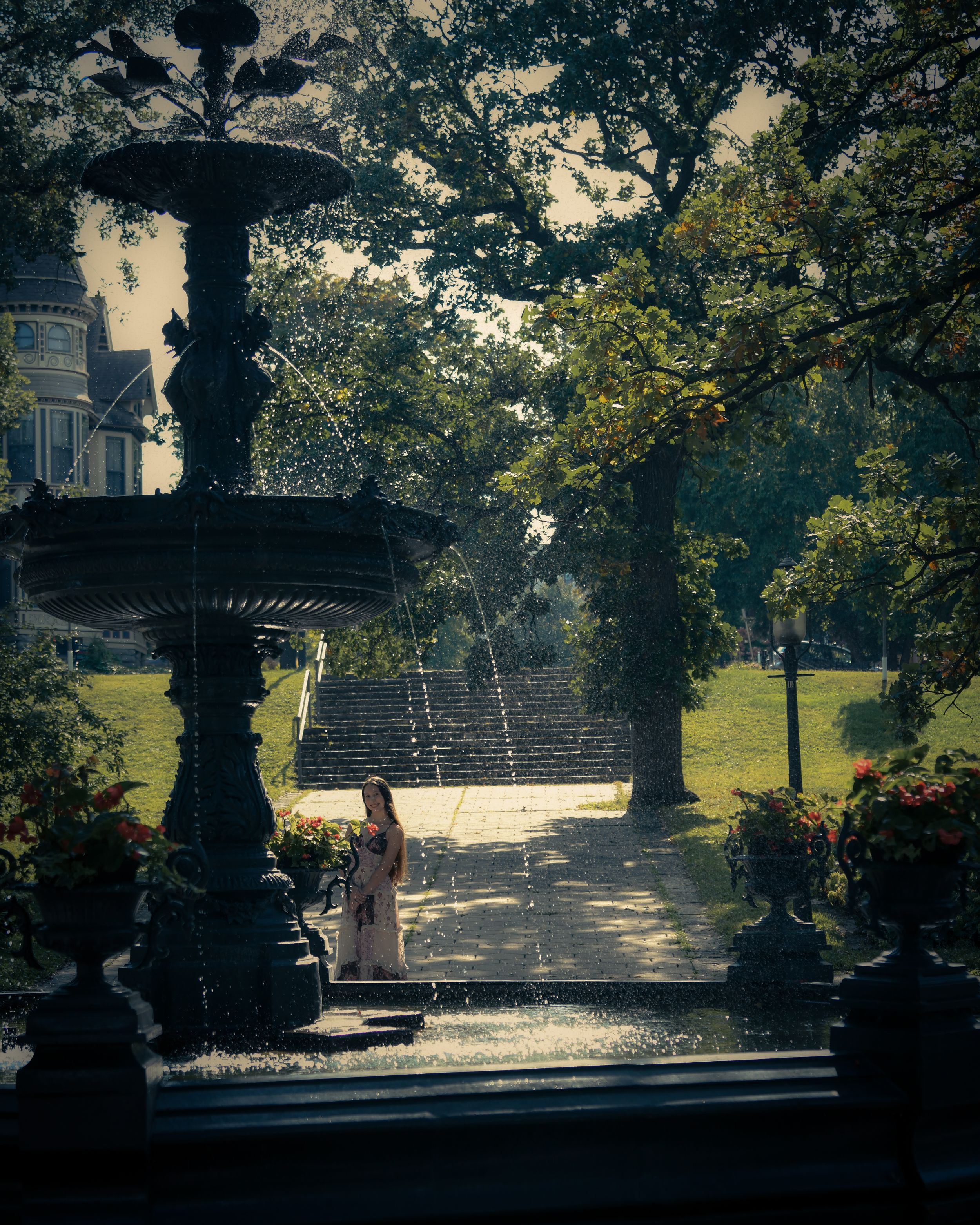 Woman standing near an ornate fountain in a lush park with trees and a historic building in the background.