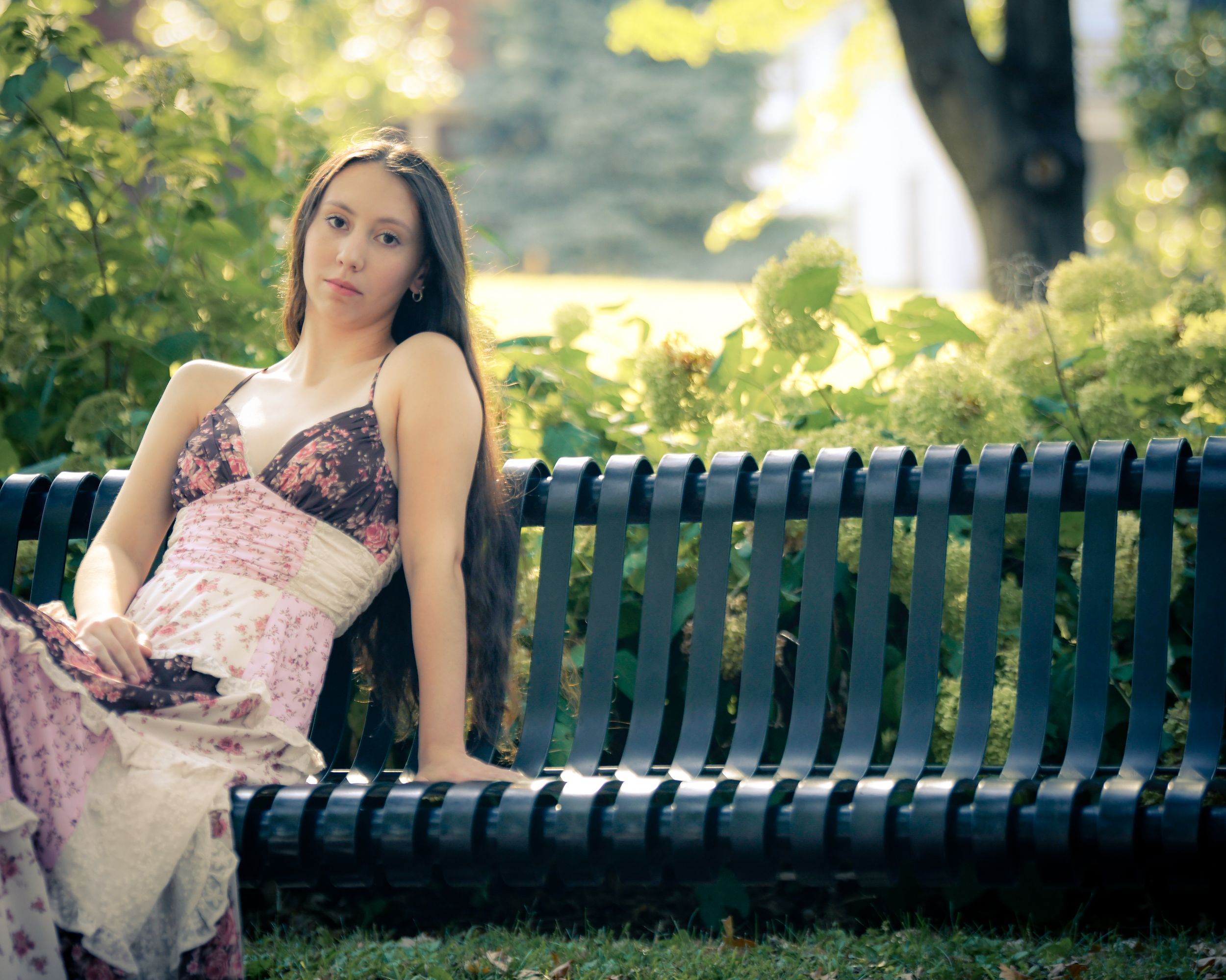 Young woman in floral dress sitting gracefully on a black park bench surrounded by lush green trees.