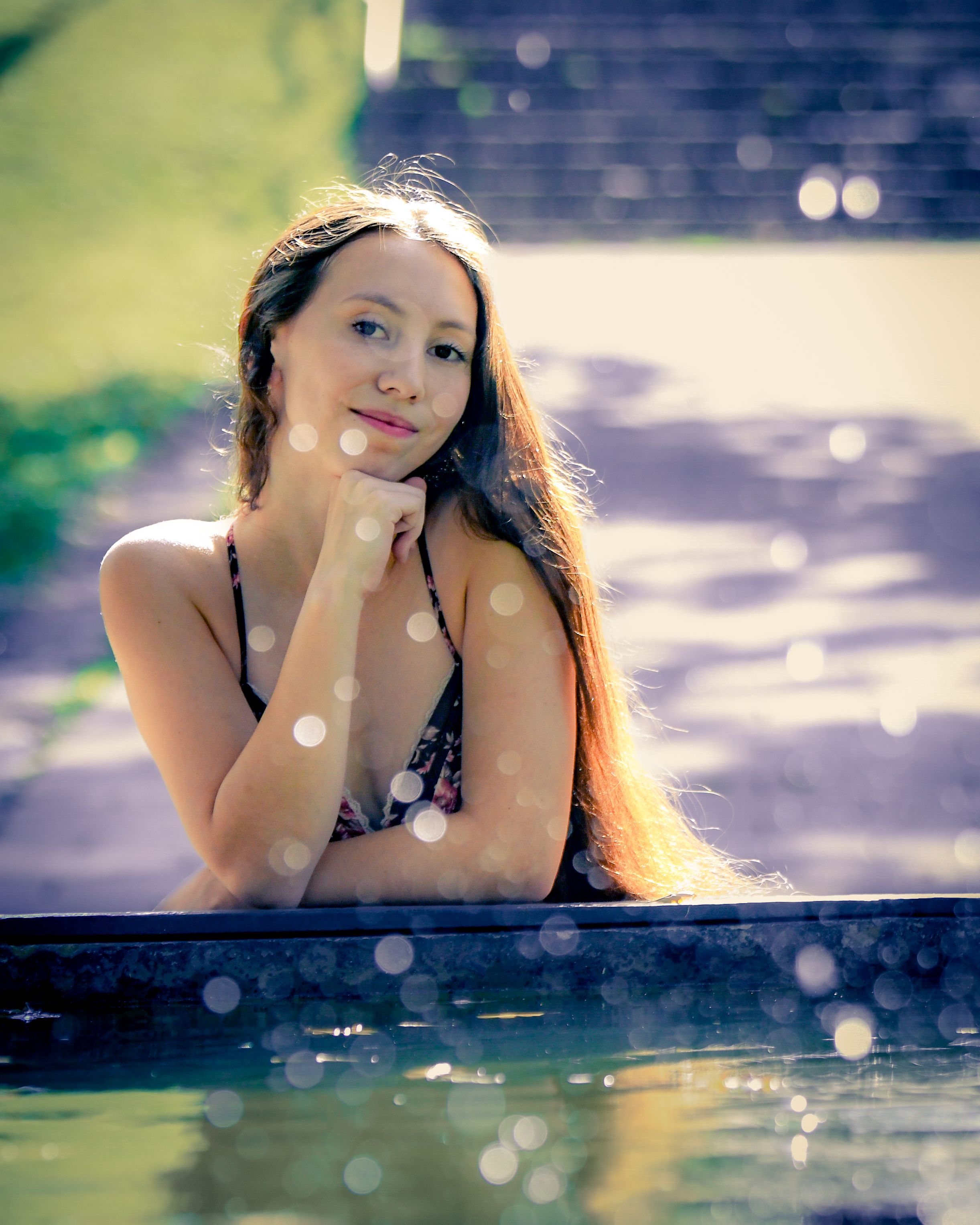 Young woman with long hair posing thoughtfully by a pool, wearing a floral bikini top in soft bokeh sunlight.