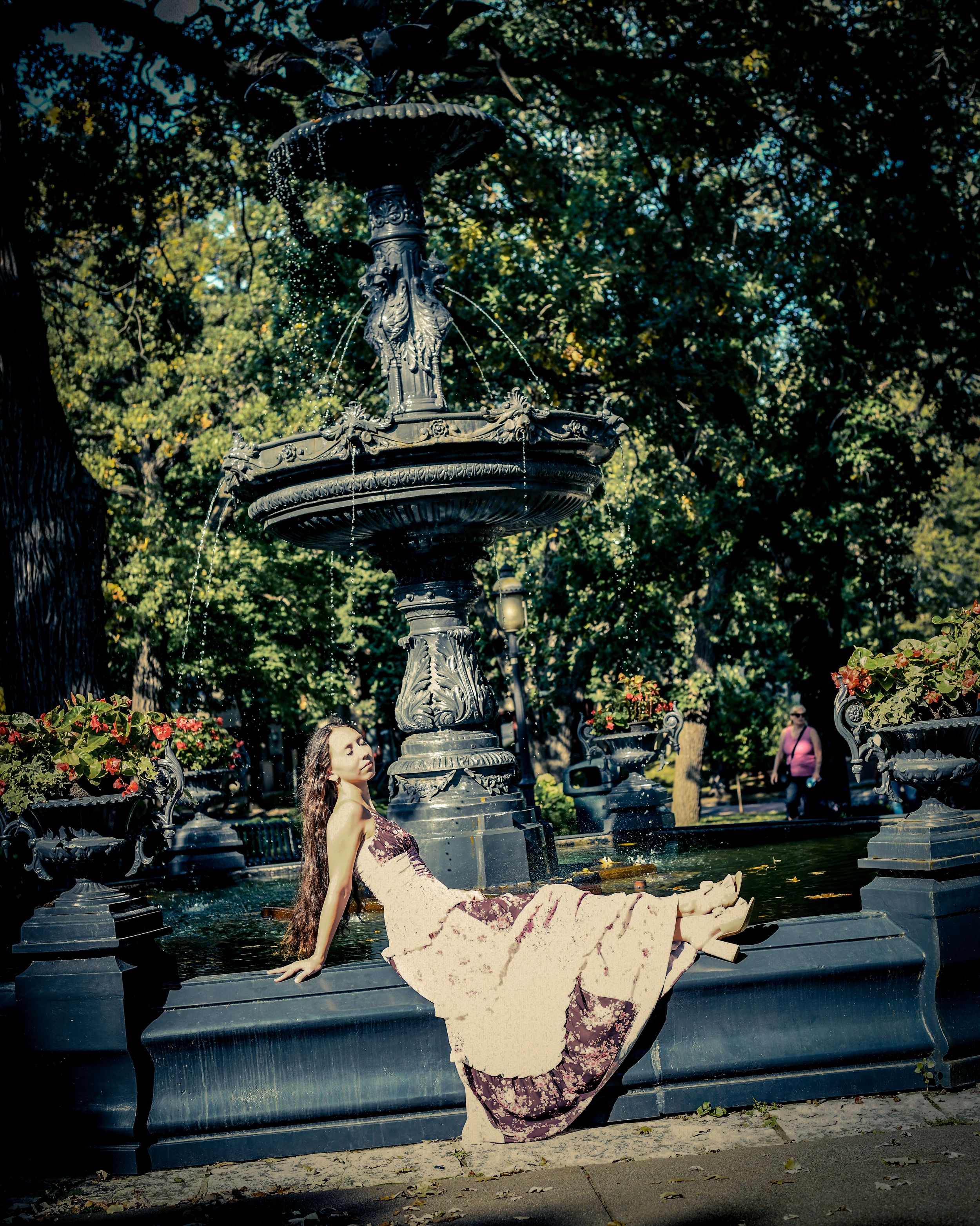Woman in flowing gown poses gracefully by ornate iron fountain surrounded by lush green trees in a park.