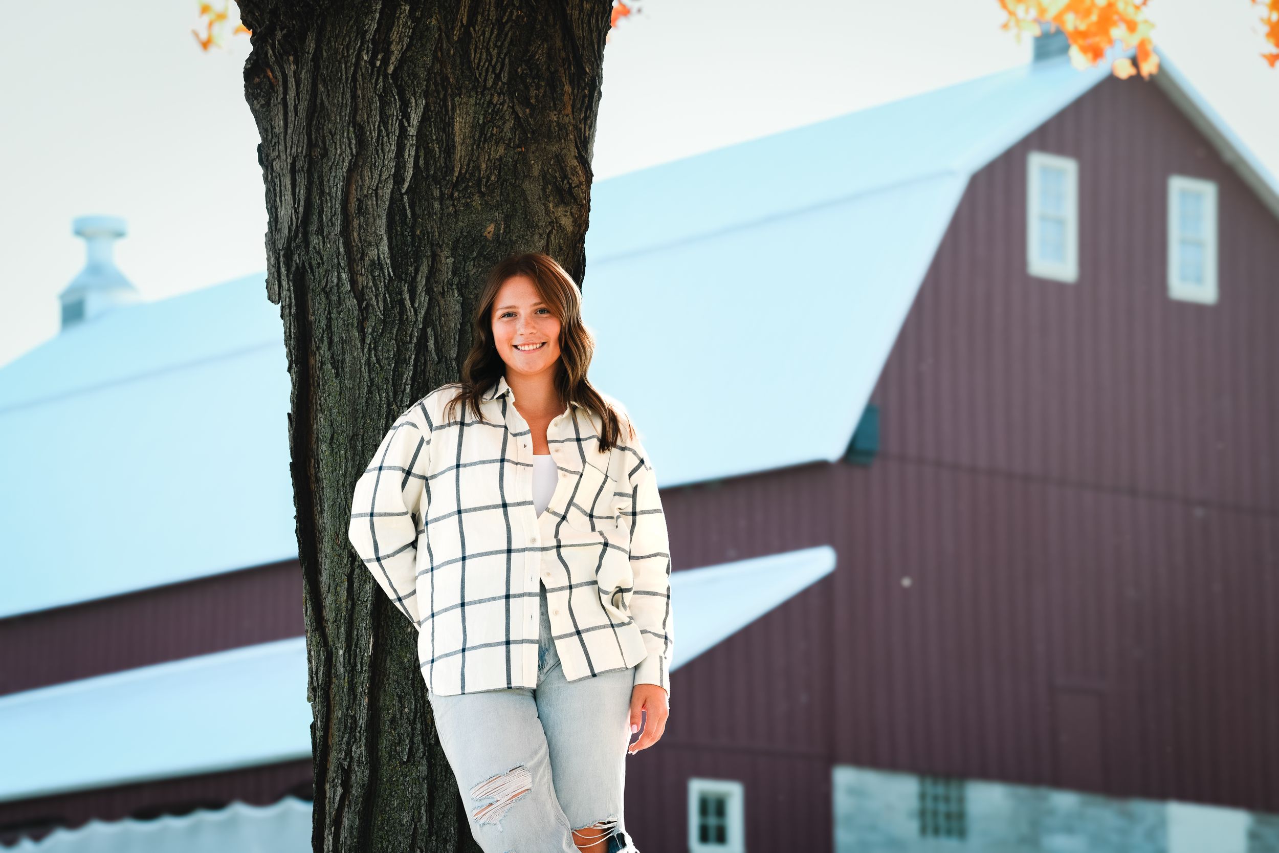 Young woman in plaid shirt leaning against tree with red barn in autumn background.