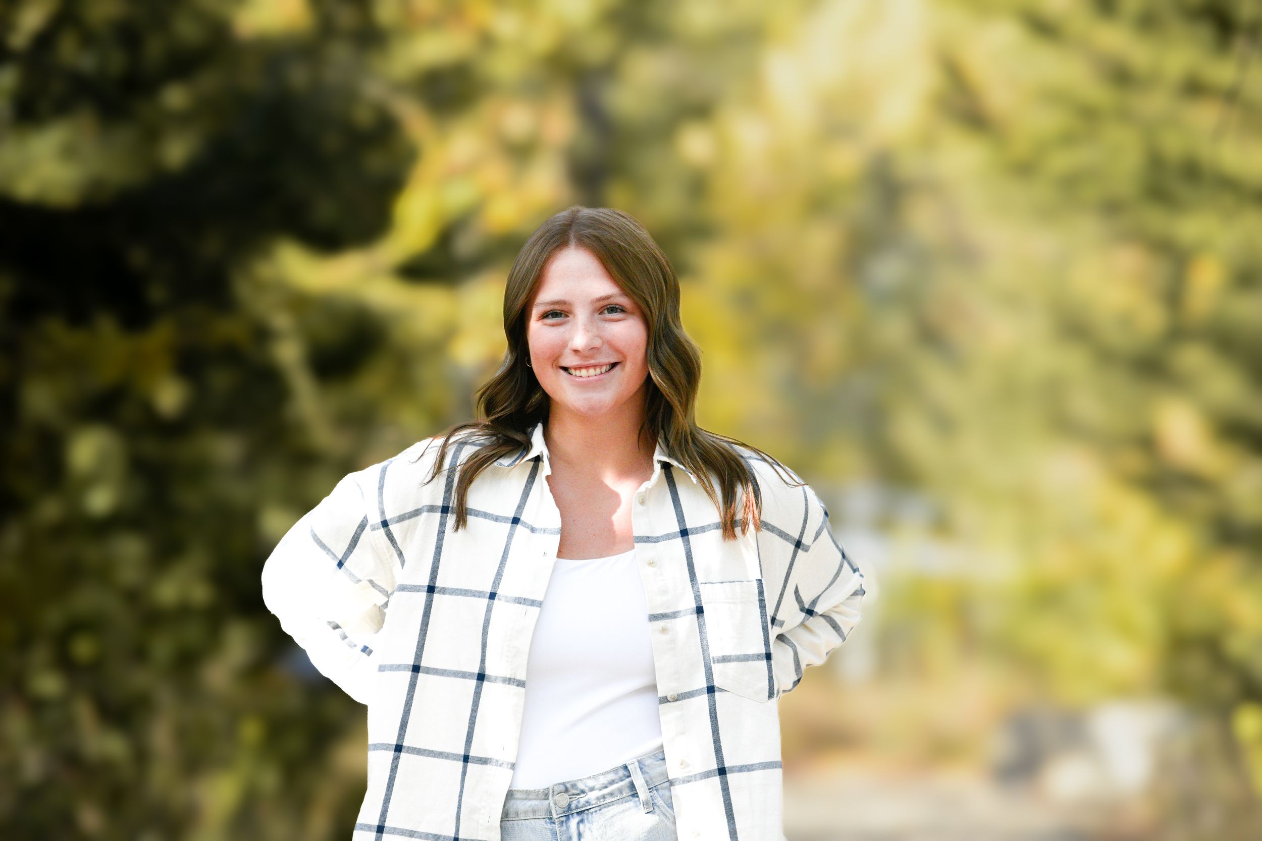 Young woman smiling outdoors in plaid shirt and jeans, posing with hands on hips among autumn trees.